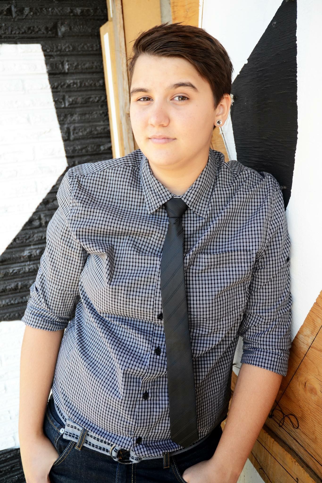 Person with short hair wearing checkered shirt and black tie leaning against white brick wall.