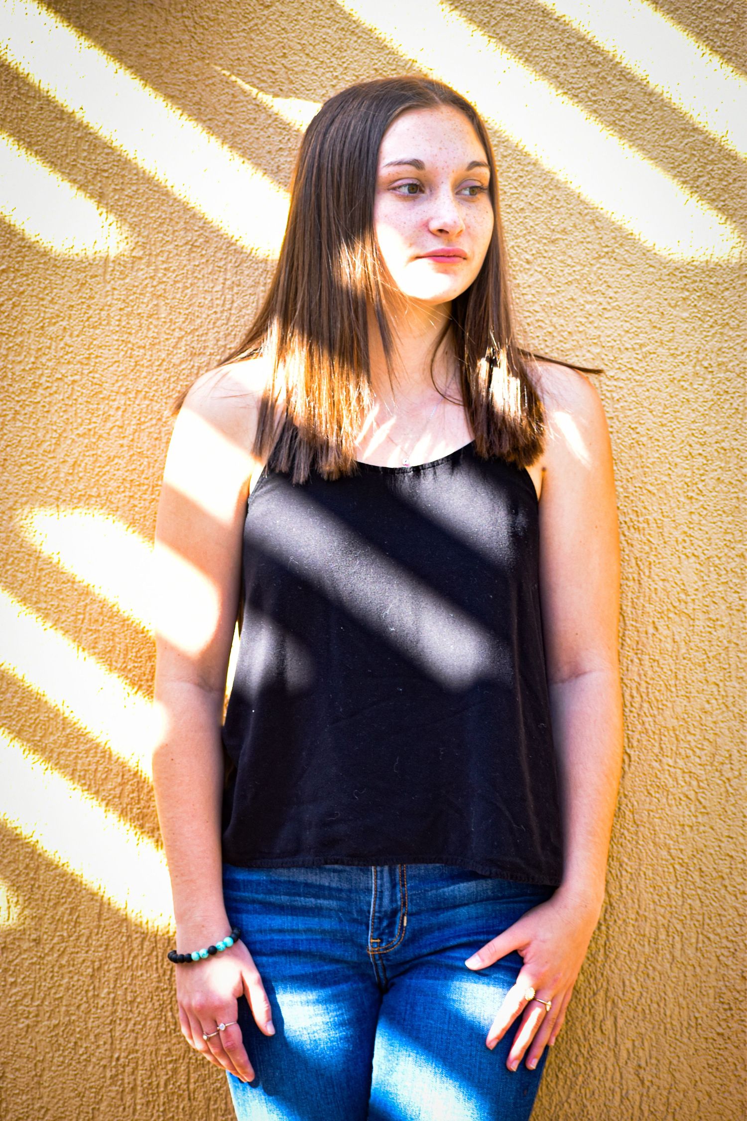 Teen girl in black tank top and jeans leans against a textured golden wall in sunlight.