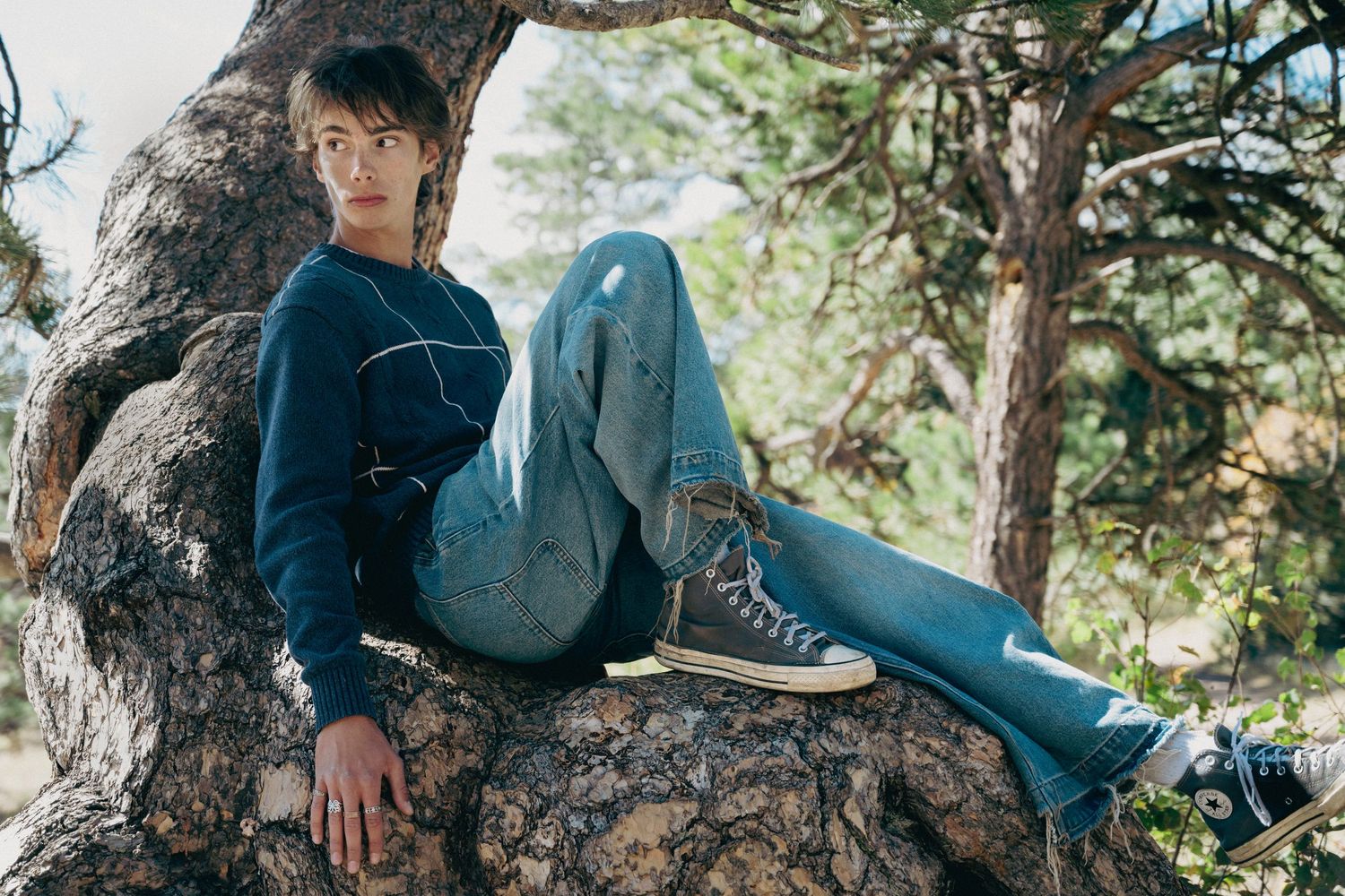 Teen boy in navy sweater and jeans sitting relaxed on large tree root in forest.