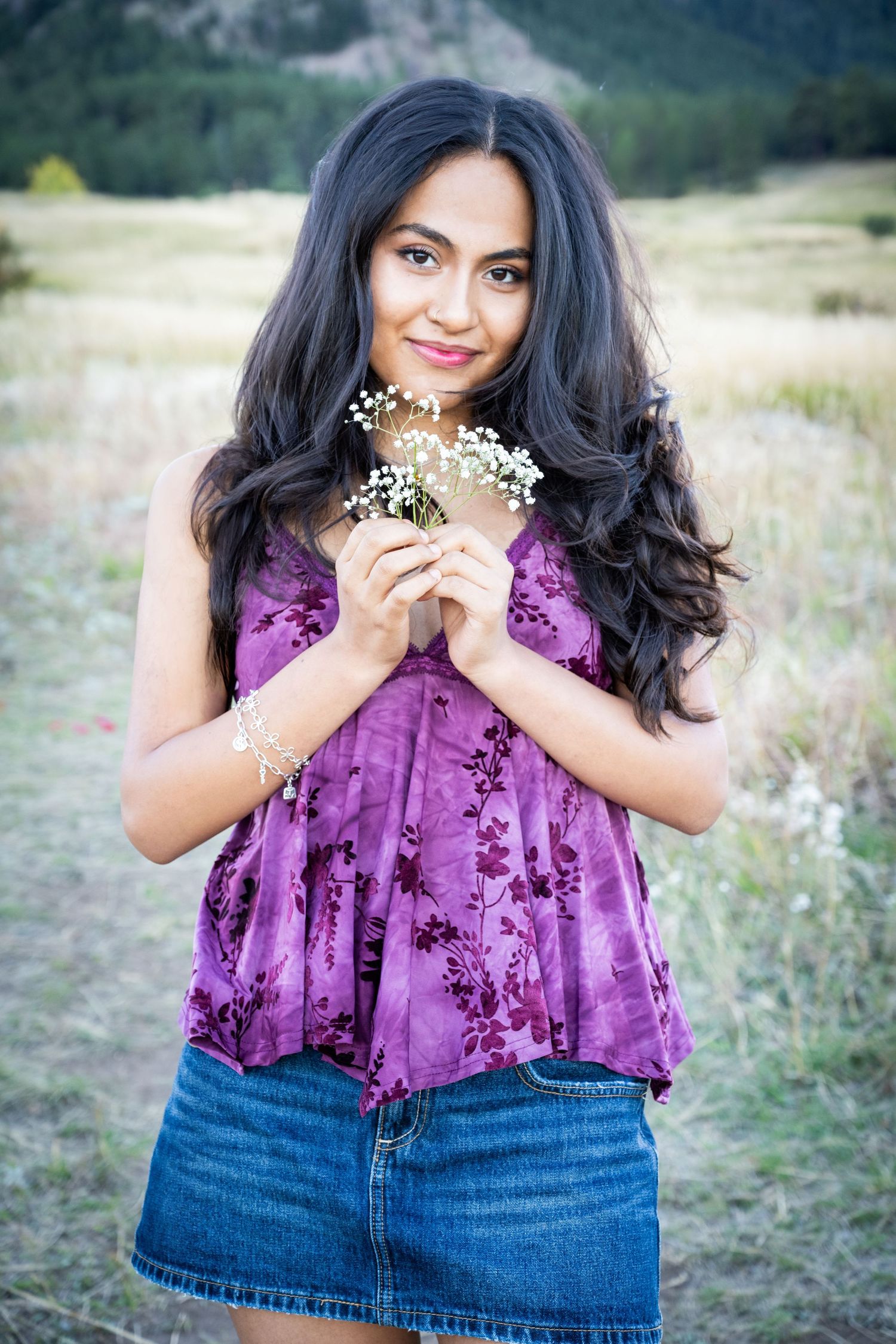 Young woman in purple floral top holding white wildflowers in mountain meadow setting.