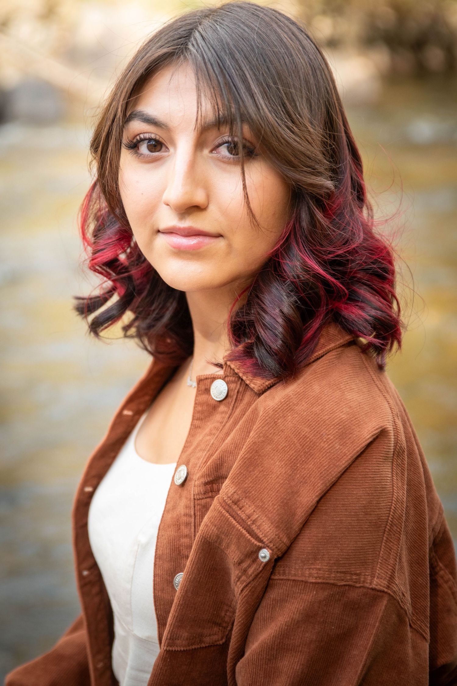 Young woman with red-highlighted brown hair wearing brown corduroy jacket near a stream.