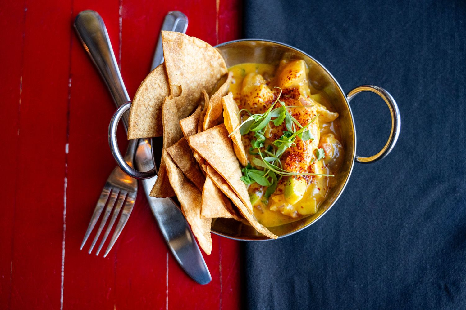 Overhead view of golden curry with crispy flatbread chips in a brass bowl on a red surface.