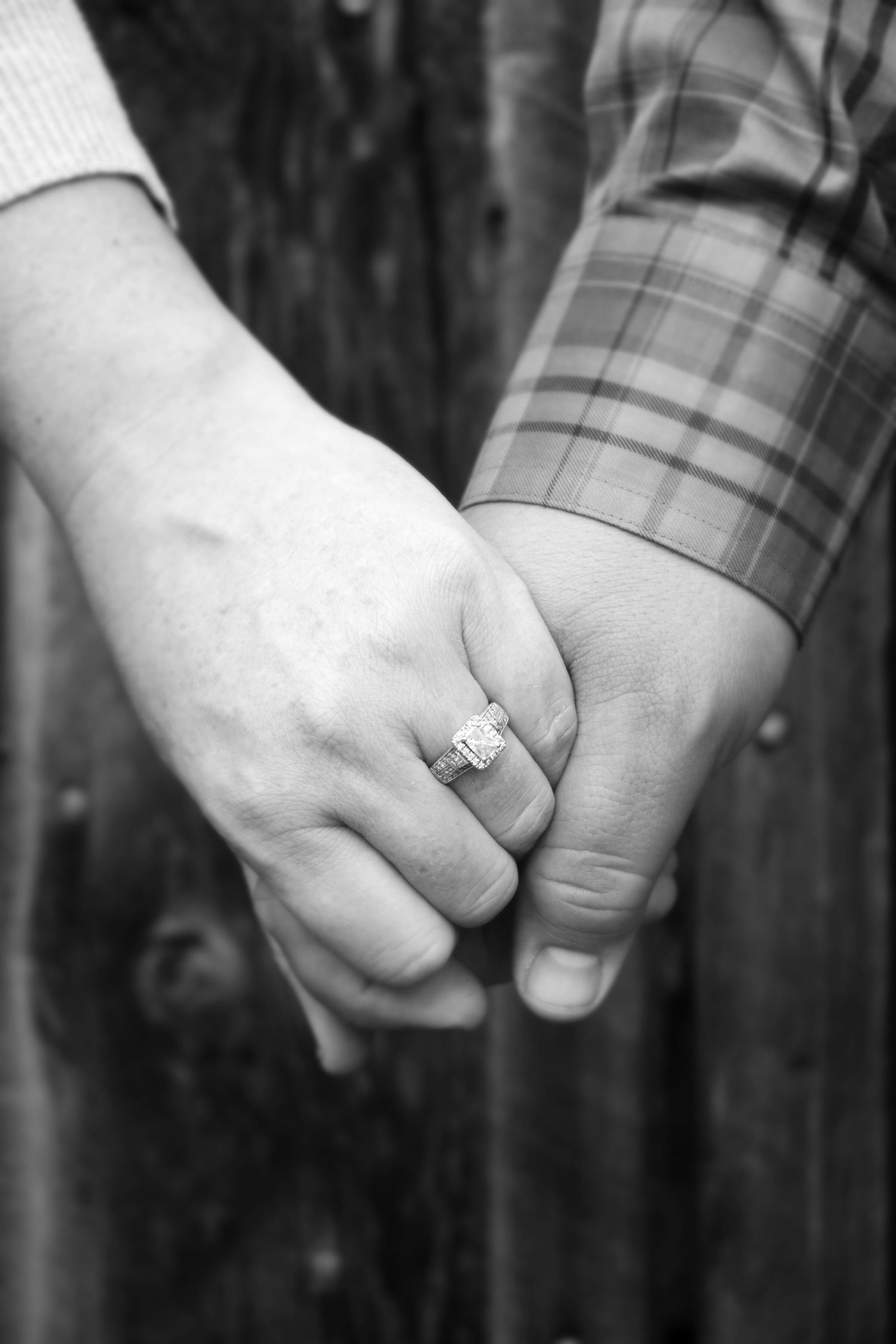 Black and white close-up of couple holding hands displaying a diamond engagement ring near a wooden fence.