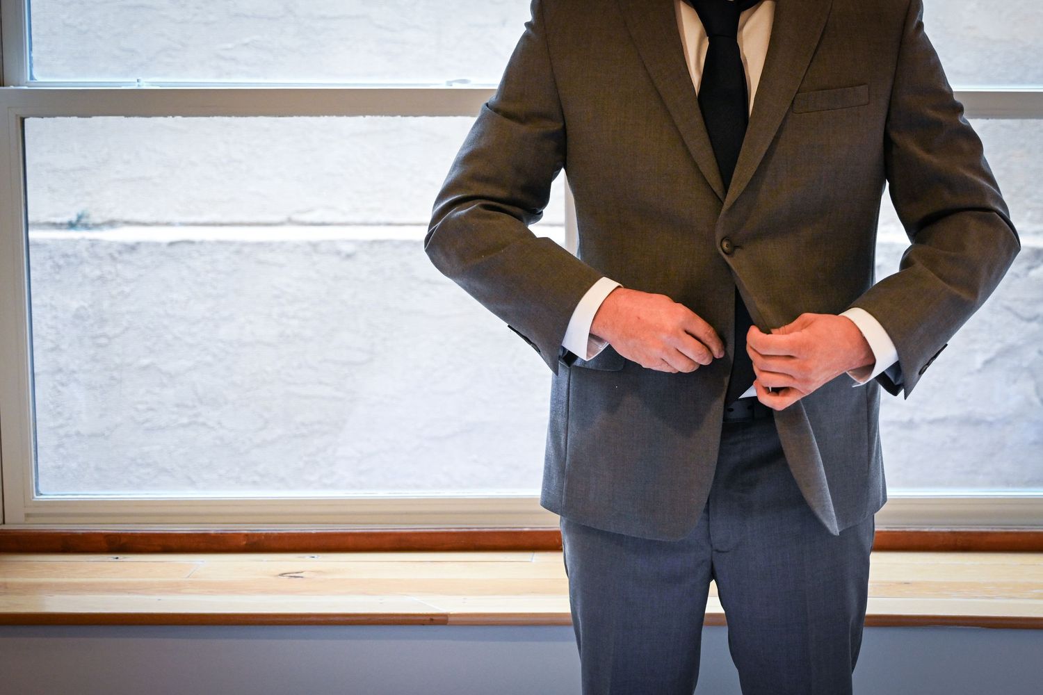Groom in gray suit buttoning jacket while standing near a bright window.