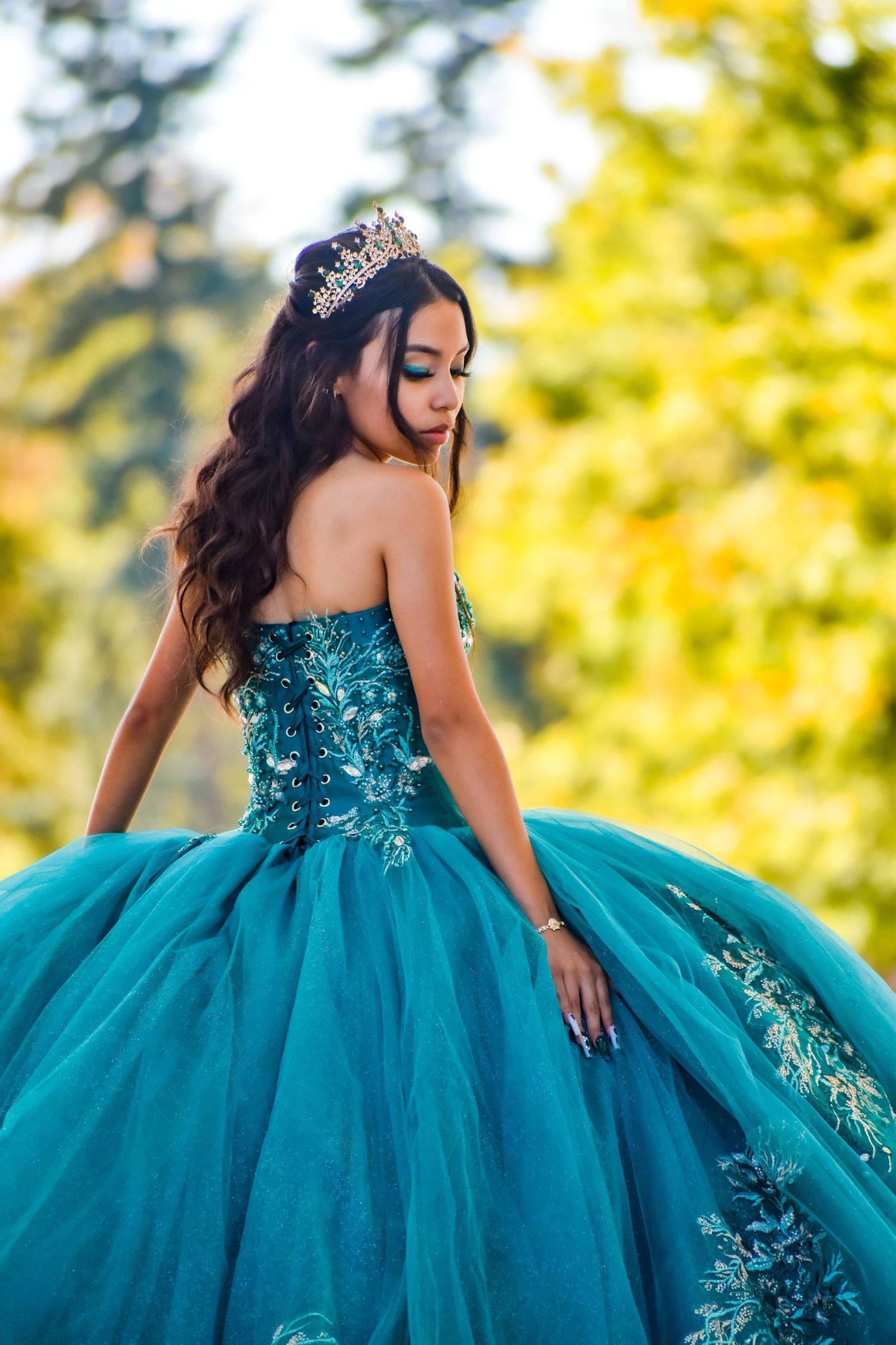 Young woman in teal ball gown with floral embroidery and crown poses outdoors among autumn foliage.