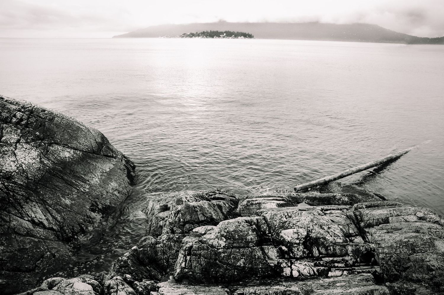 Black and white coastal landscape with rocky shoreline, calm water, and misty island in the background.