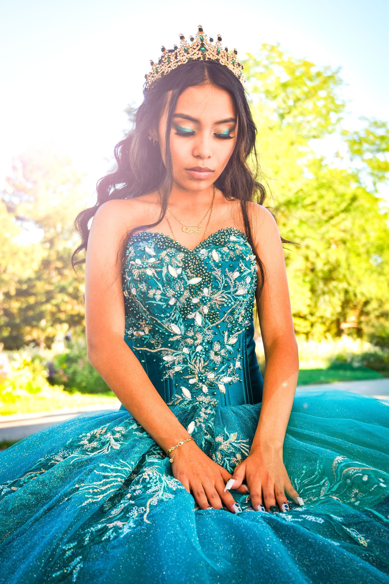 Young woman in teal quinceañera gown with gold crown and embroidered bodice poses outdoors in sunlight.