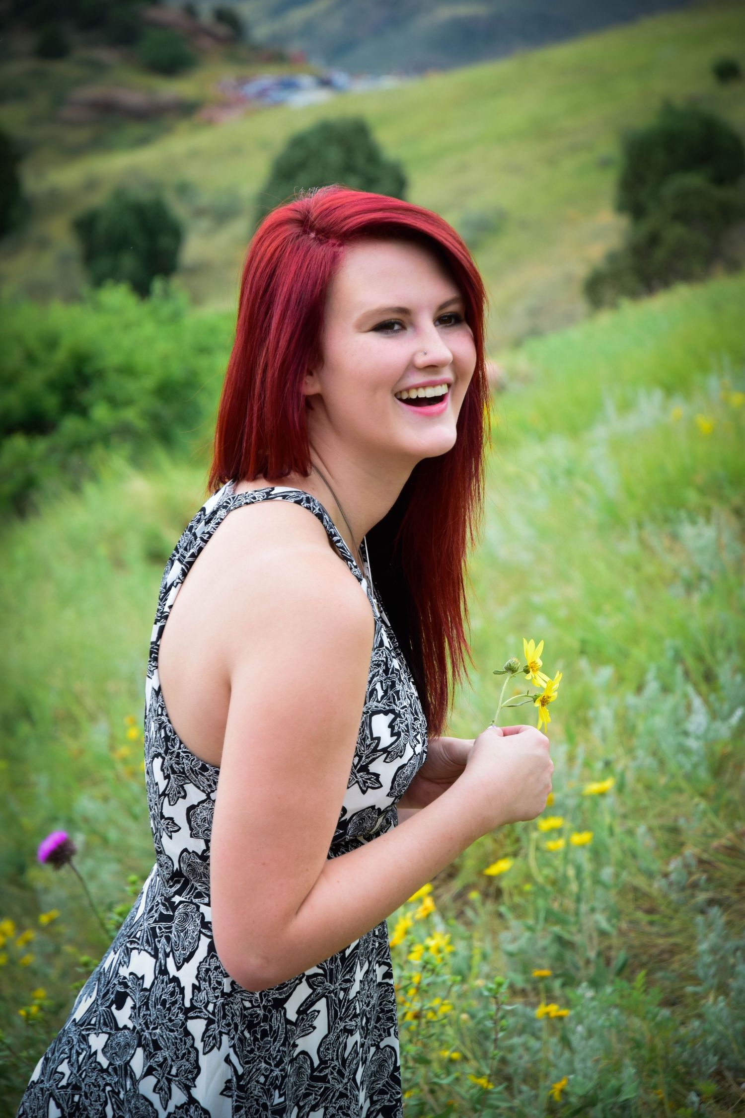 Laughing young woman with red hair holding wildflowers in a lush green meadow.