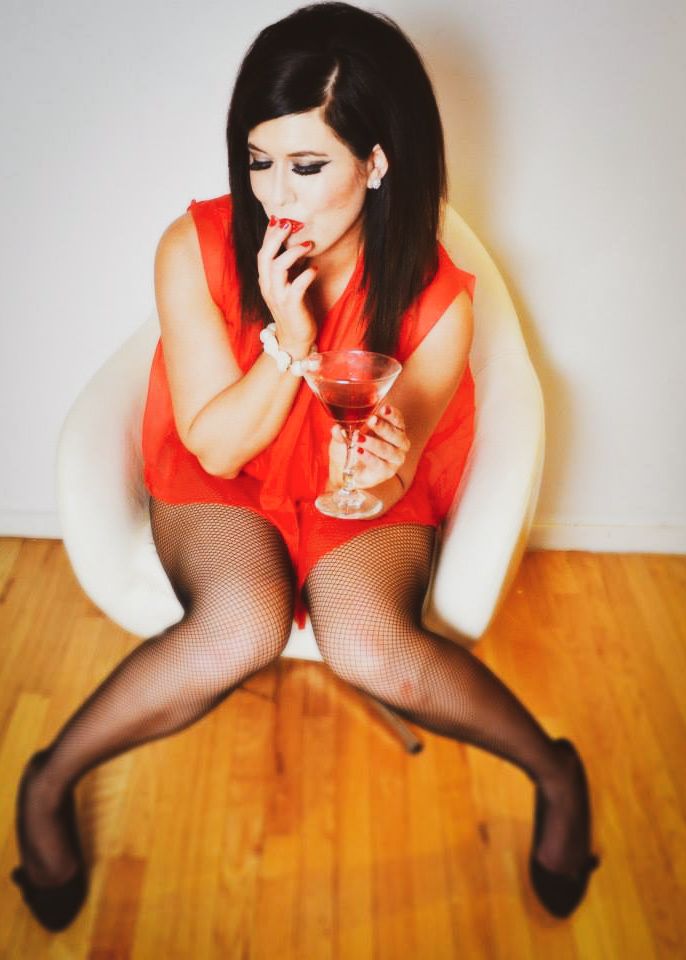 Woman in red dress and fishnet stockings sitting in white chair holding a red cocktail drink.