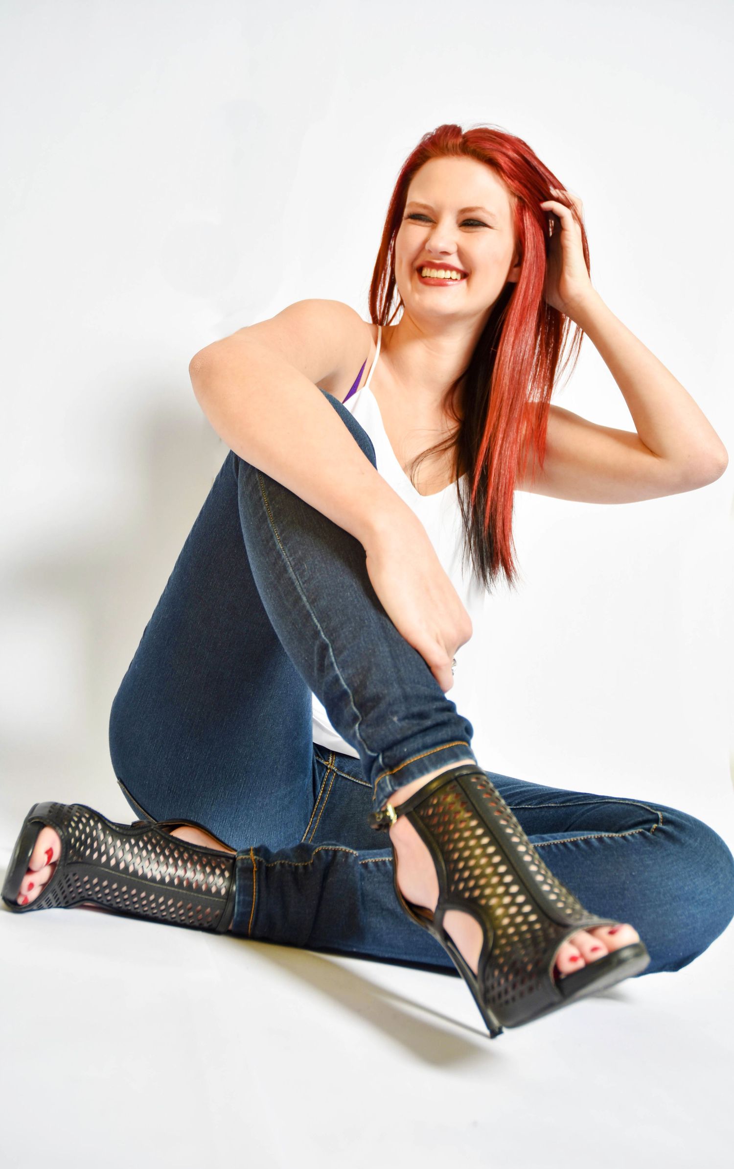 Smiling redhead woman sitting on white studio floor wearing blue jeans, white tank top, and black mesh boots.