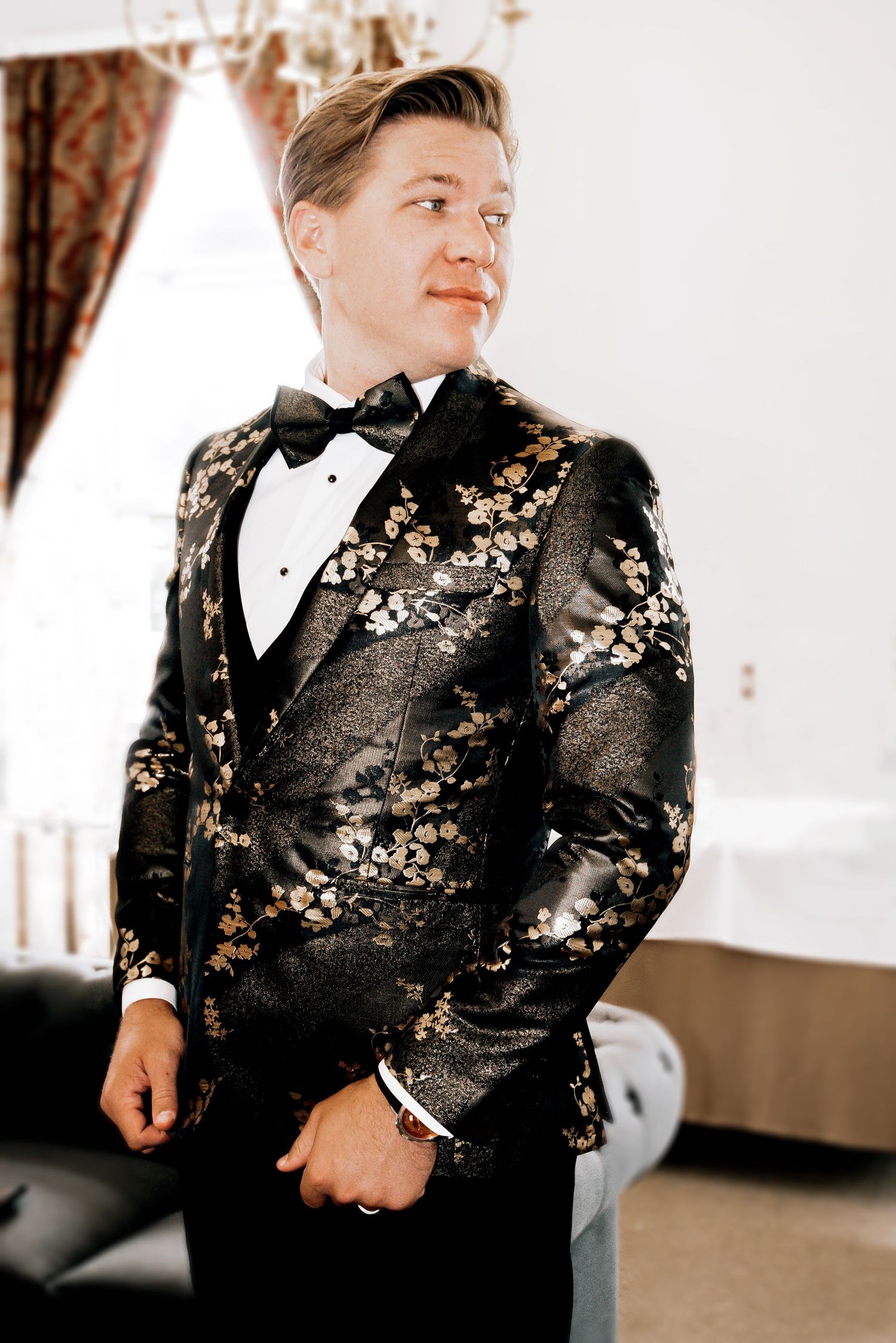 Groom poses in elegant black and gold floral tuxedo jacket with bow tie at wedding venue.