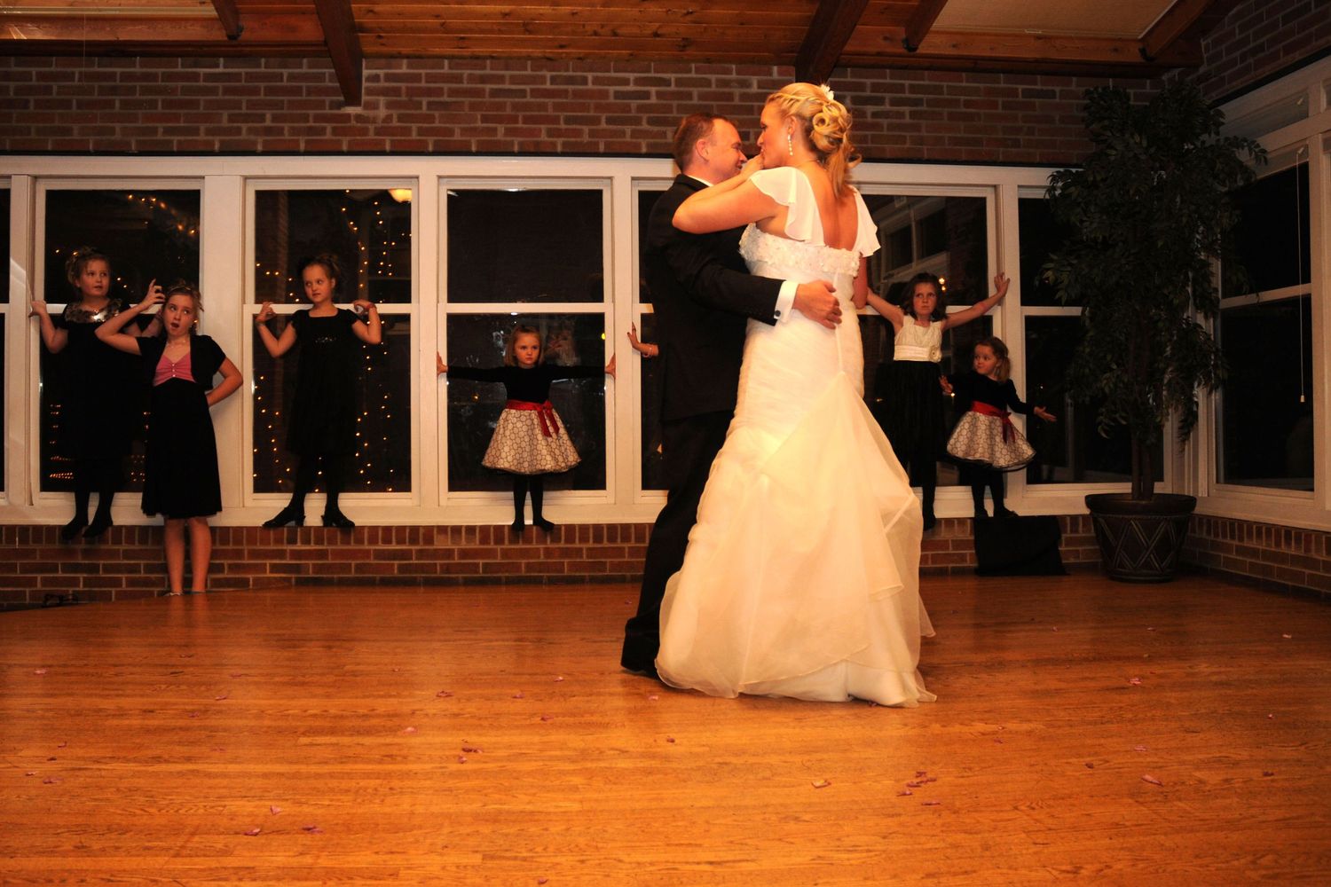 Bride and groom share their first dance on a hardwood floor while wedding guests watch in the background.