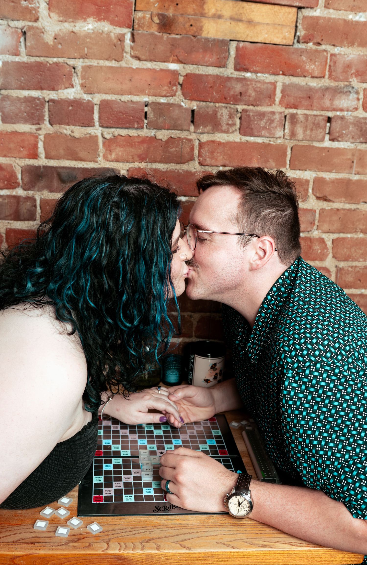 Couple sharing a kiss over a mosaic board game table against a red brick wall.