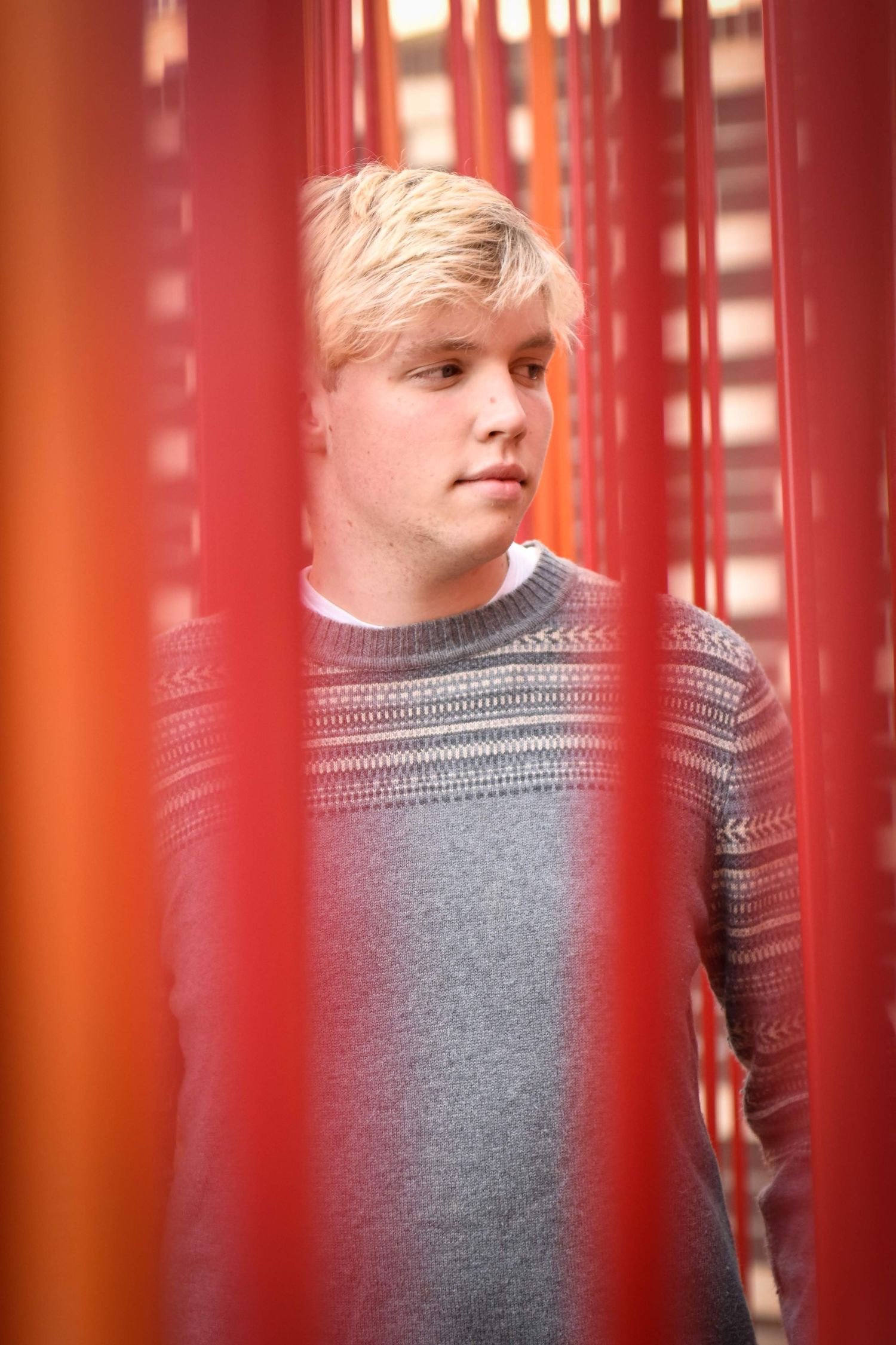 Blonde young man in gray patterned sweater seen through red vertical bars or curtains.