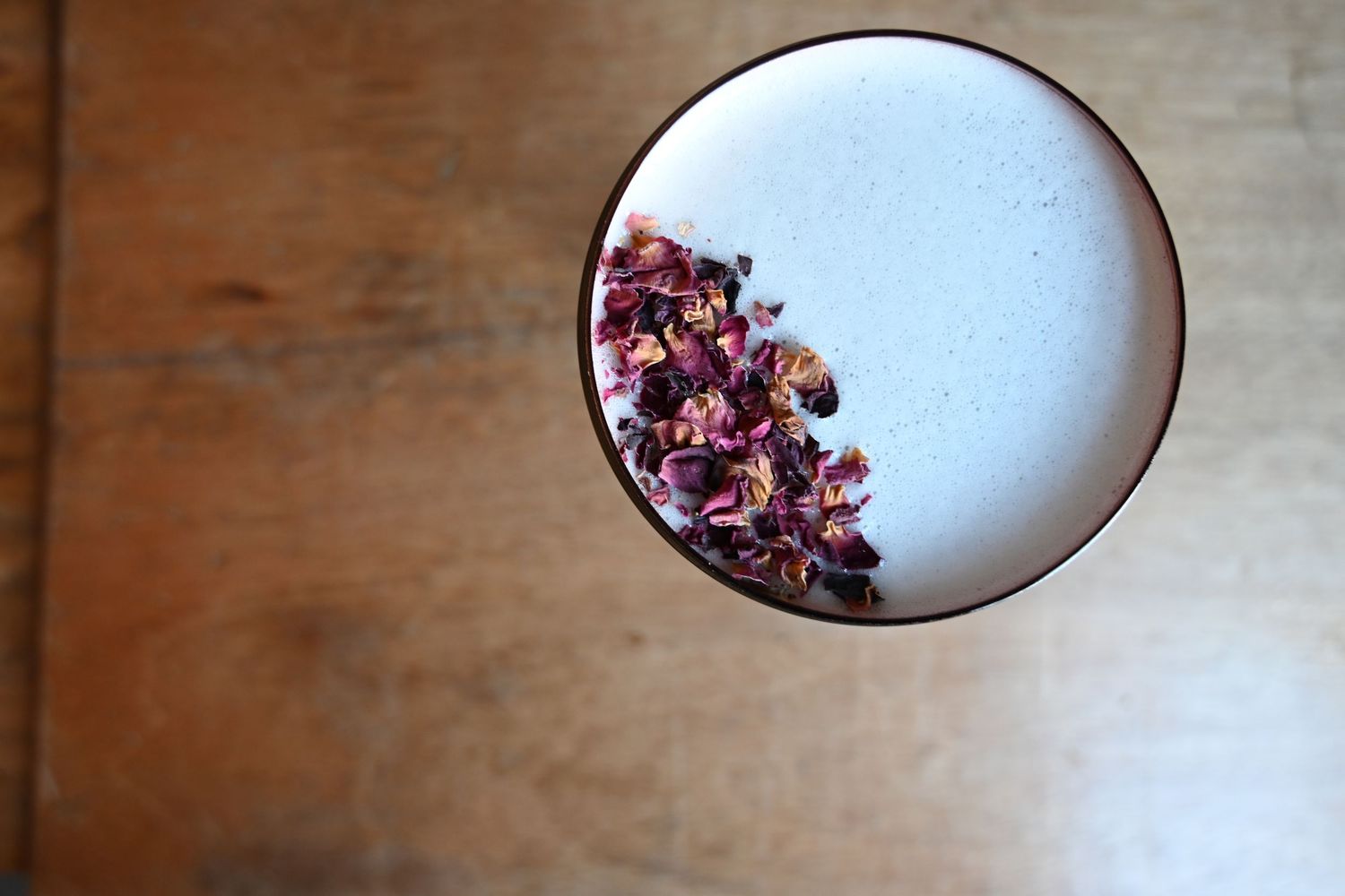 Top view of a white cocktail garnished with dried rose petals in a ceramic glass on wood.