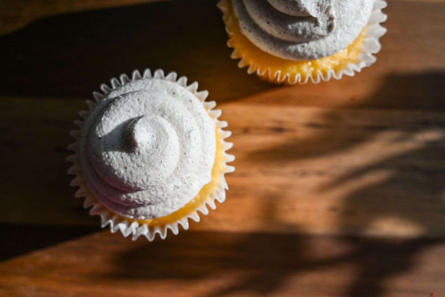 Two vanilla cupcakes with swirled gray frosting in white paper liners on a wooden surface.