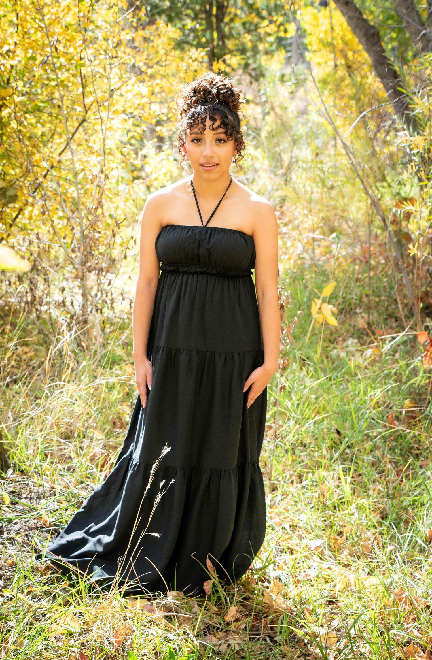 Young woman in elegant black halter maxi dress stands among golden autumn foliage outdoors.