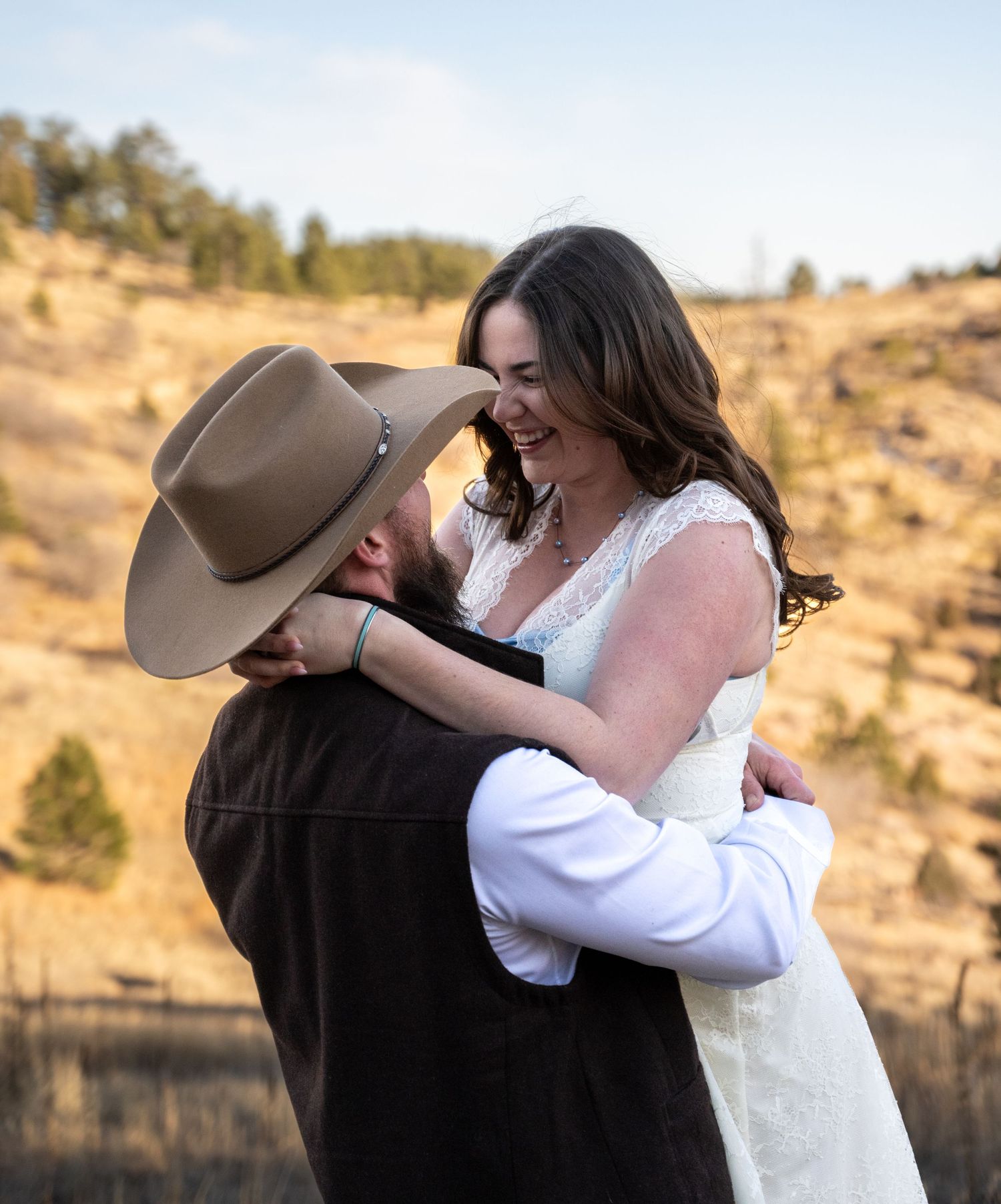 Happy bride in white dress laughing as groom in cowboy hat lifts her during outdoor wedding celebration.