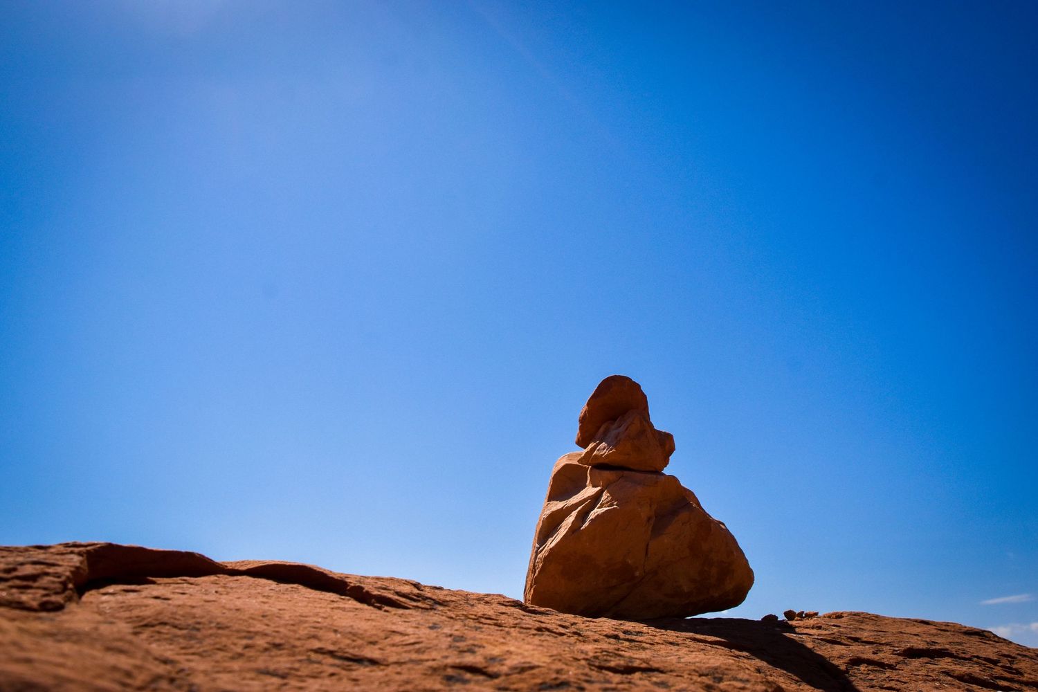 A balanced rock cairn sits atop red sandstone against a vivid blue sky in a desert landscape.