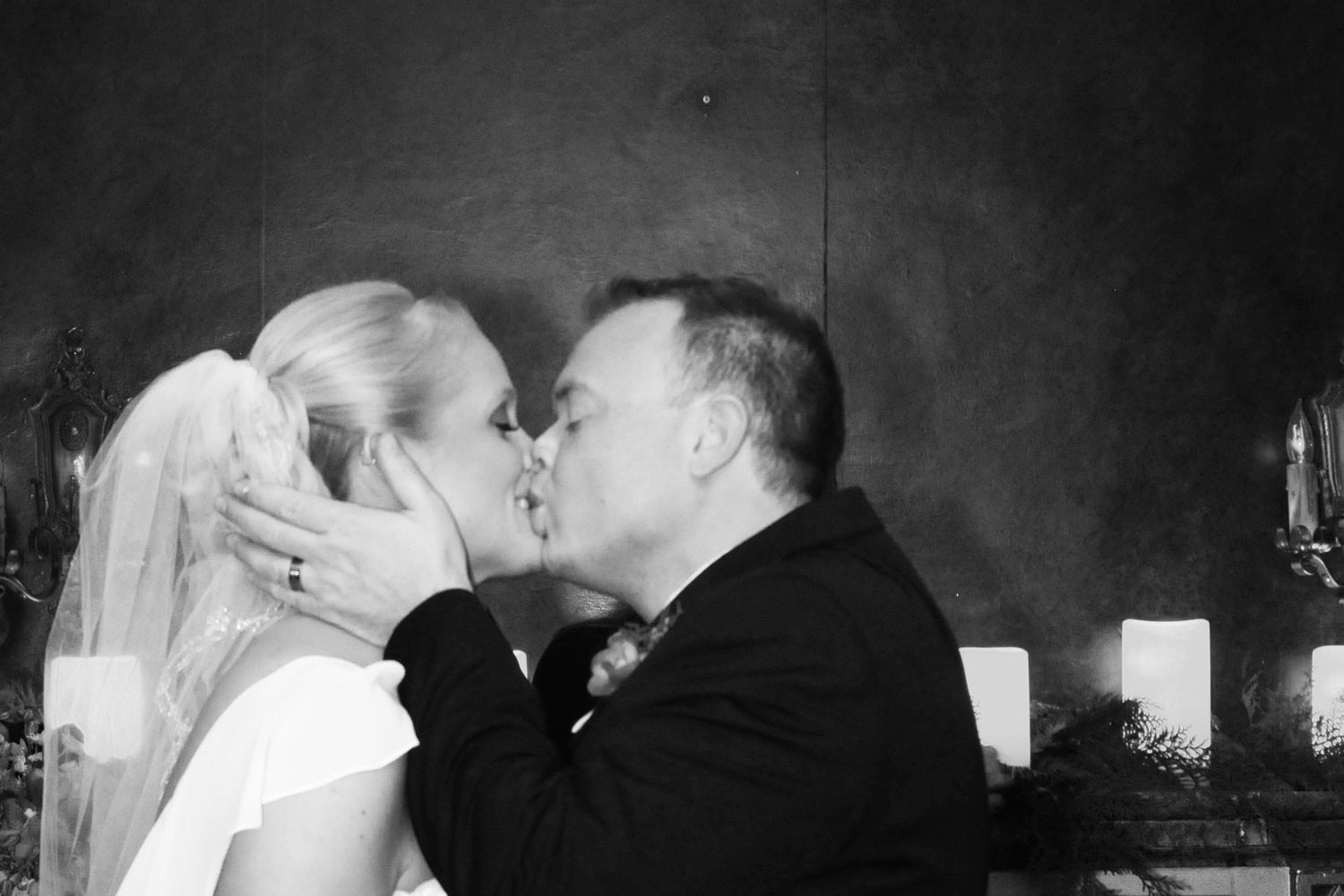 Black and white photo of a bride and groom sharing their first kiss at the altar with candles behind them.