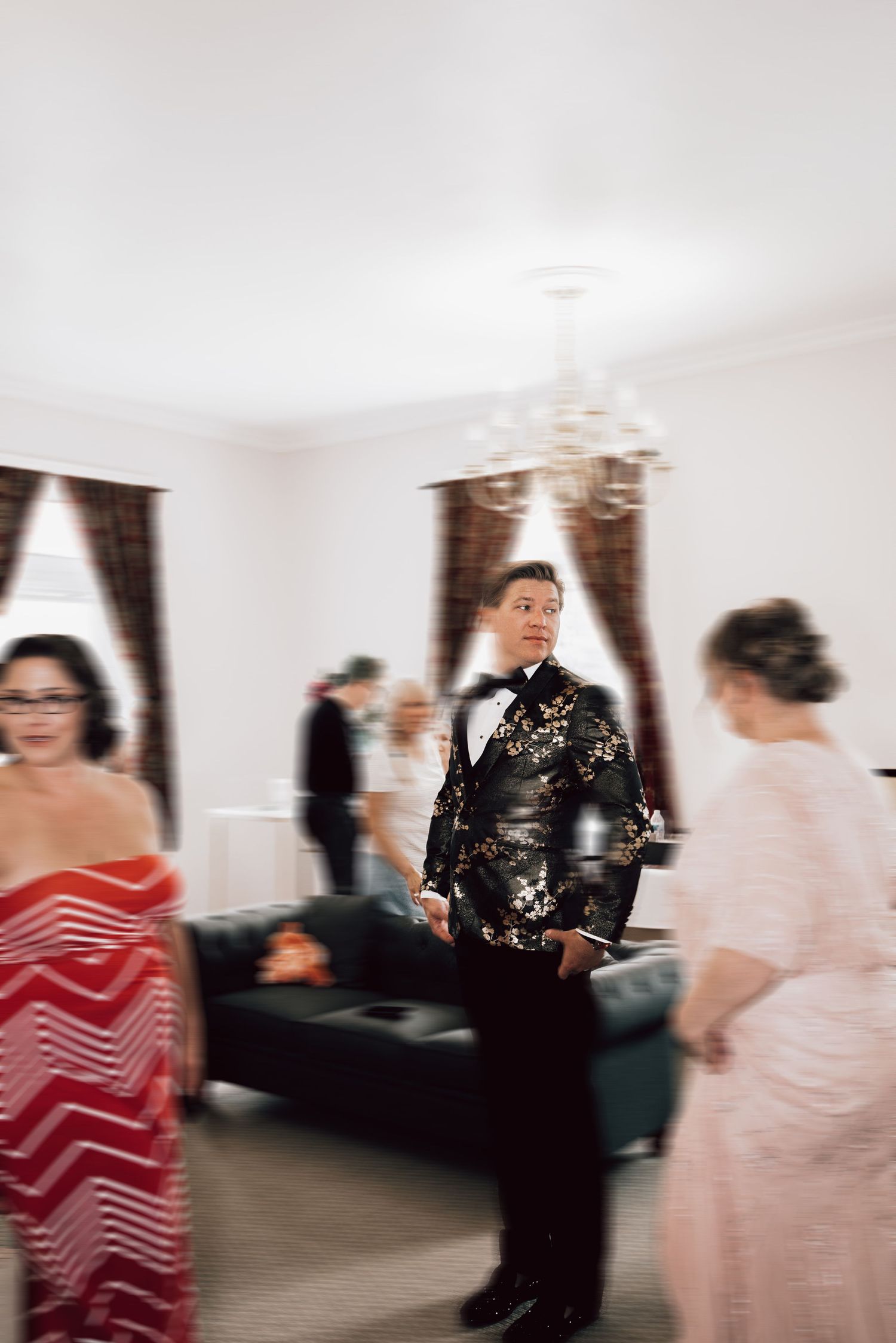 Groom in black and gold floral tuxedo mingles with wedding guests in elegant white reception room.