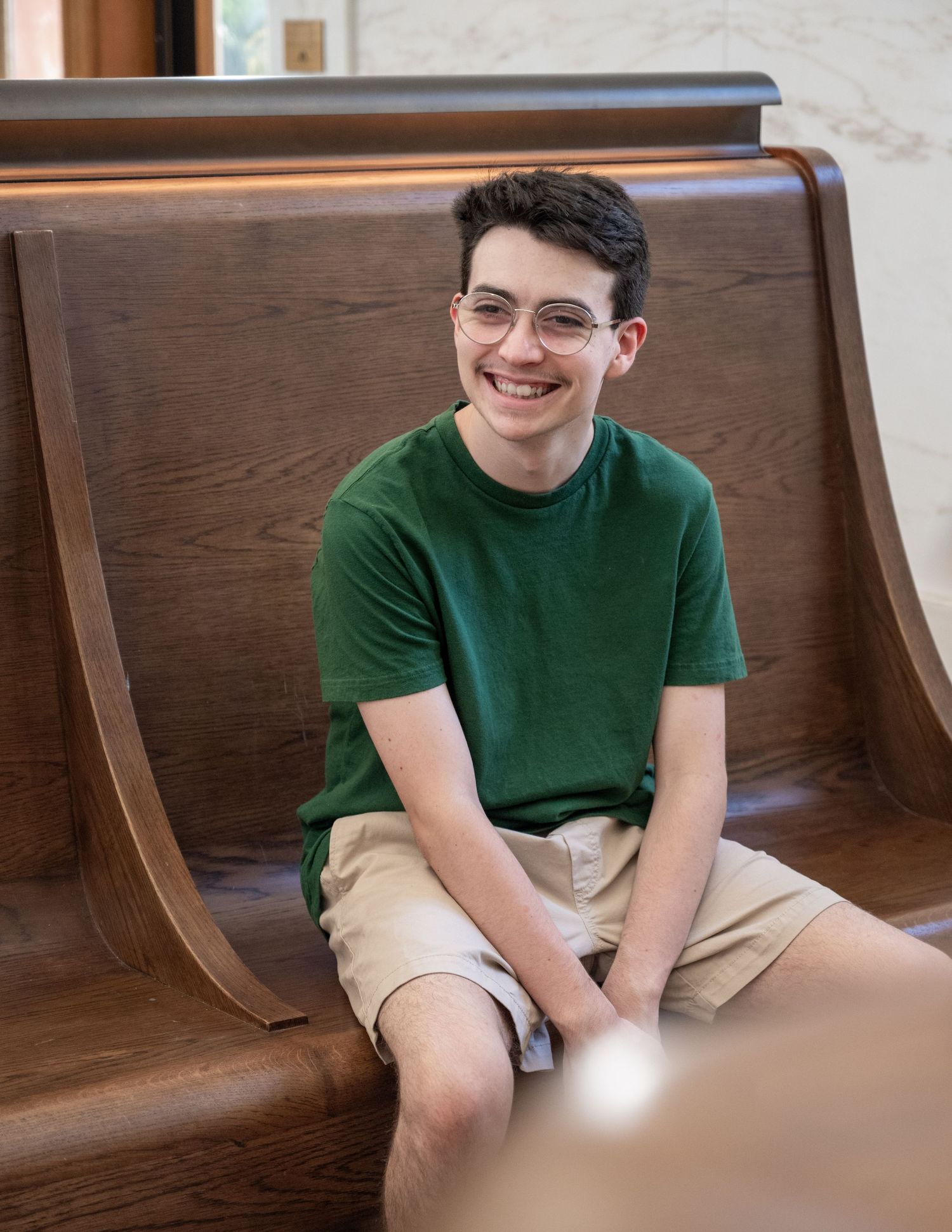 Smiling teen boy with glasses in green shirt sitting on wooden church pew.