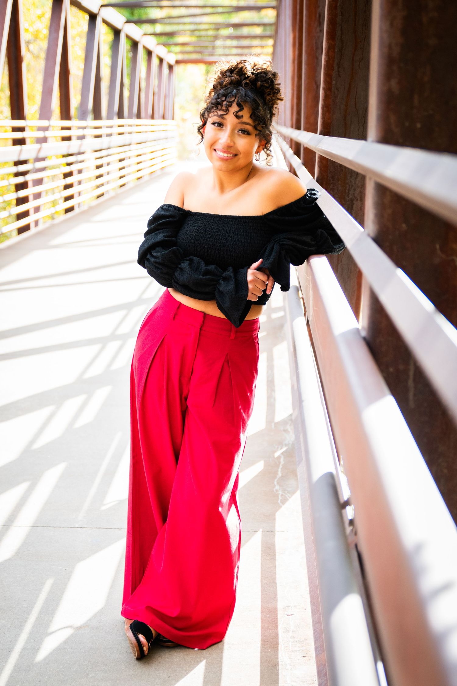 Young woman in black off-shoulder top and red wide-leg pants poses on a sunny bridge.