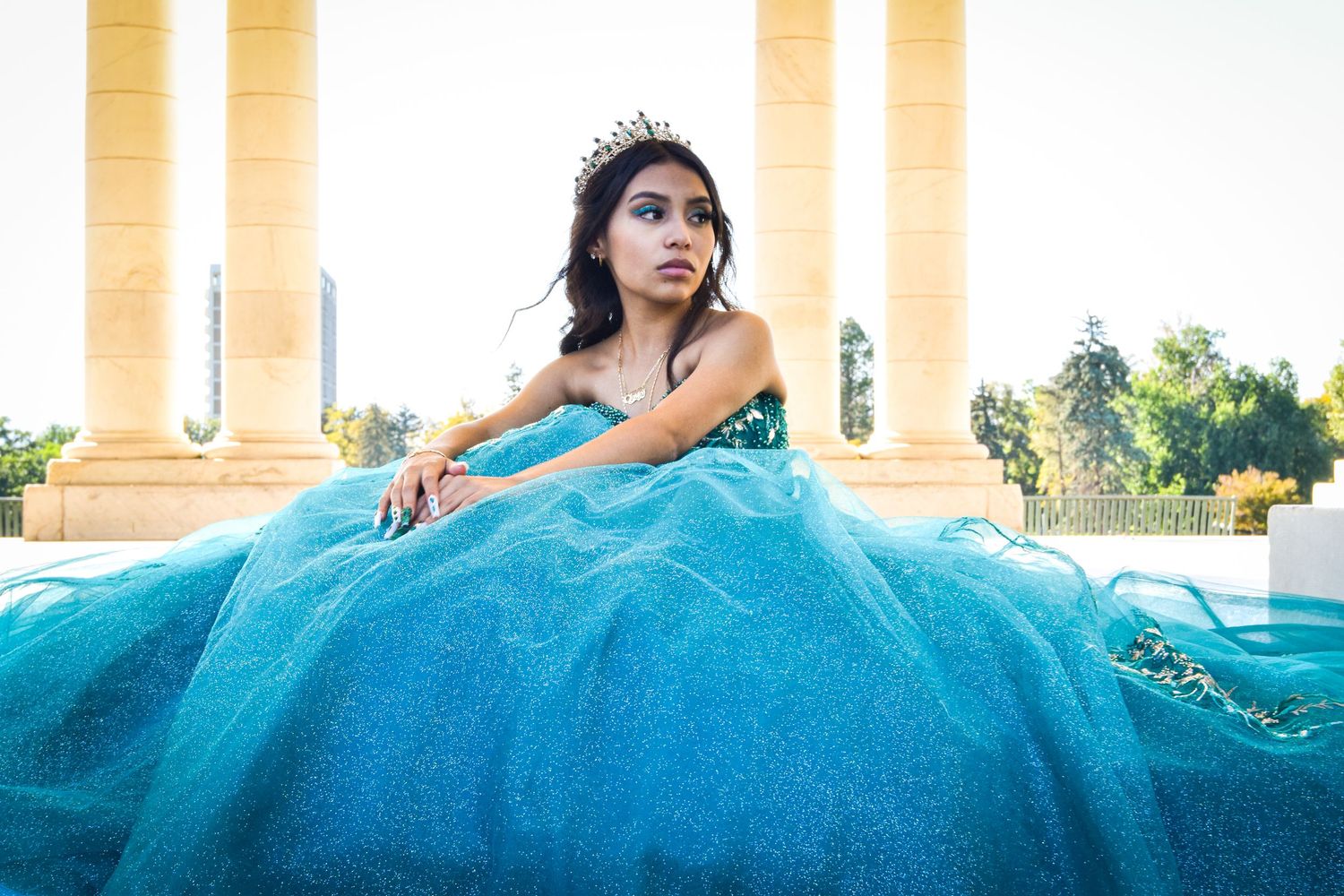 Young woman in a teal ball gown and tiara poses gracefully among stone columns at a quinceañera celebration.