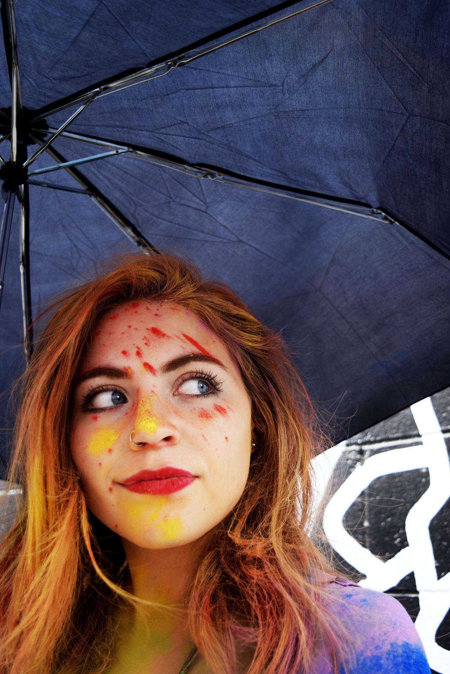 Young woman with colorful Holi paint on her face holding a dark umbrella, looking up thoughtfully.