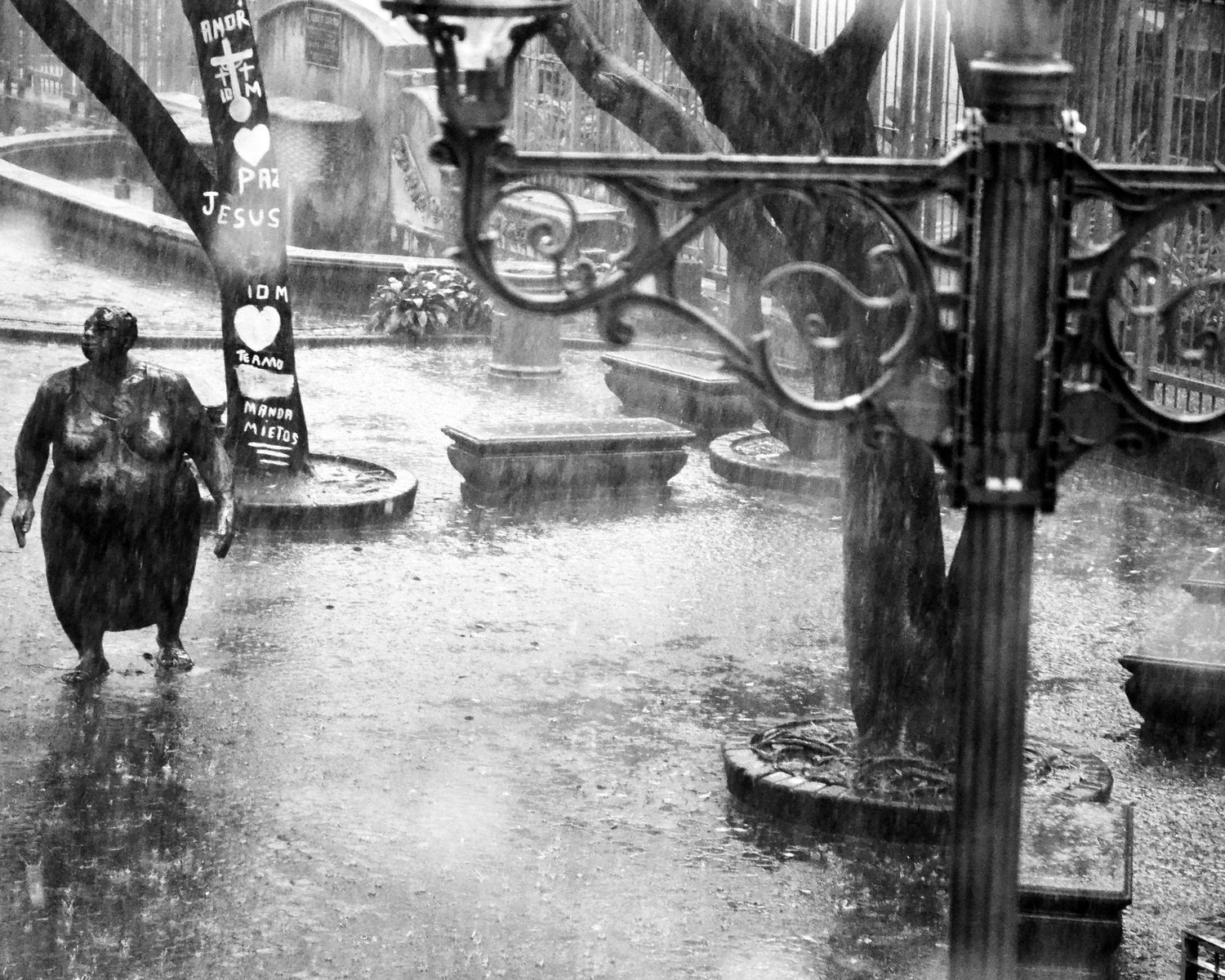 Black and white photo of a person walking in heavy rain on a wet city street with ornate iron fencing.
