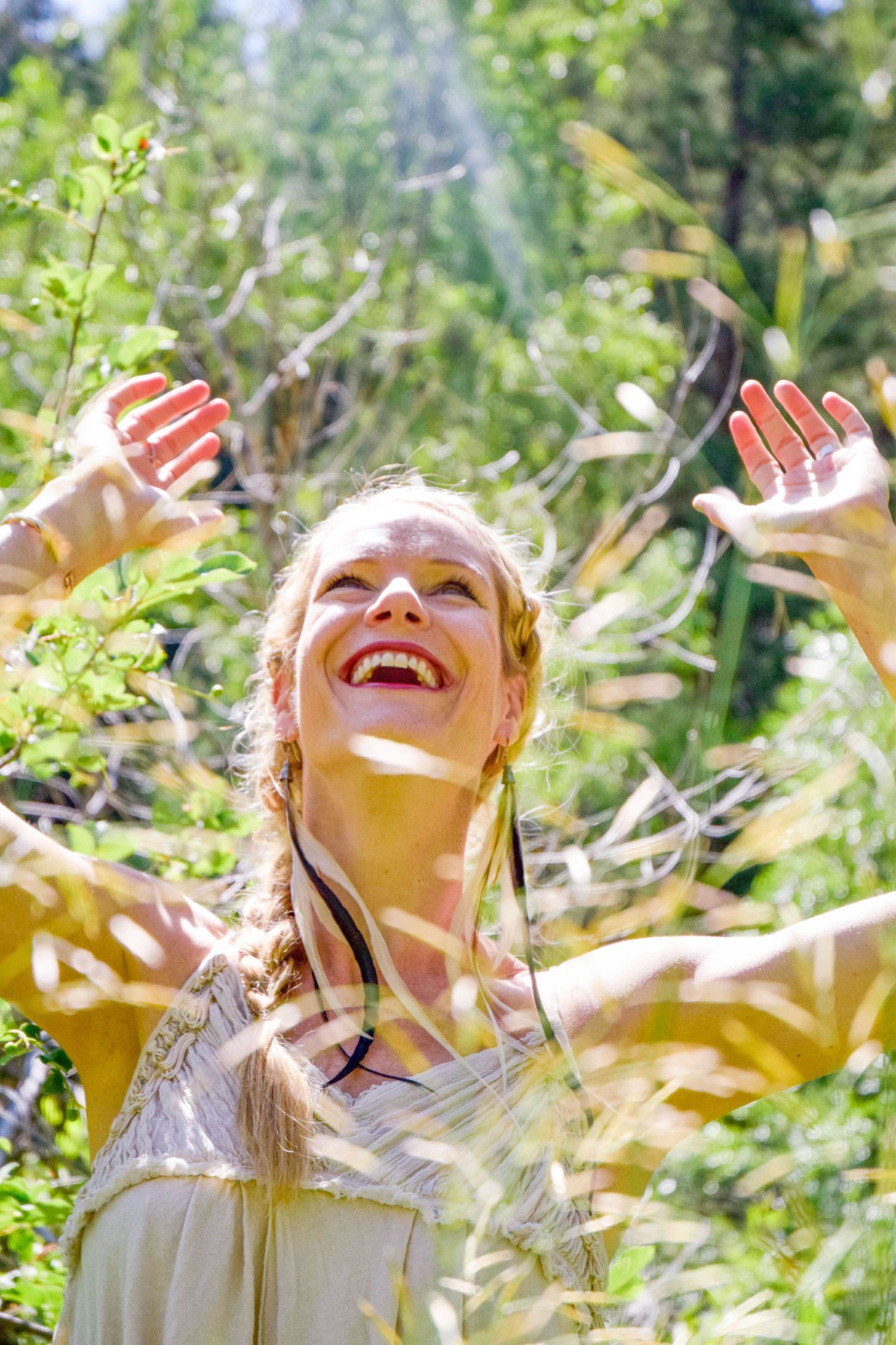 Joyful blonde woman with braids raises arms outdoors in sunlit green forest, laughing with happiness.