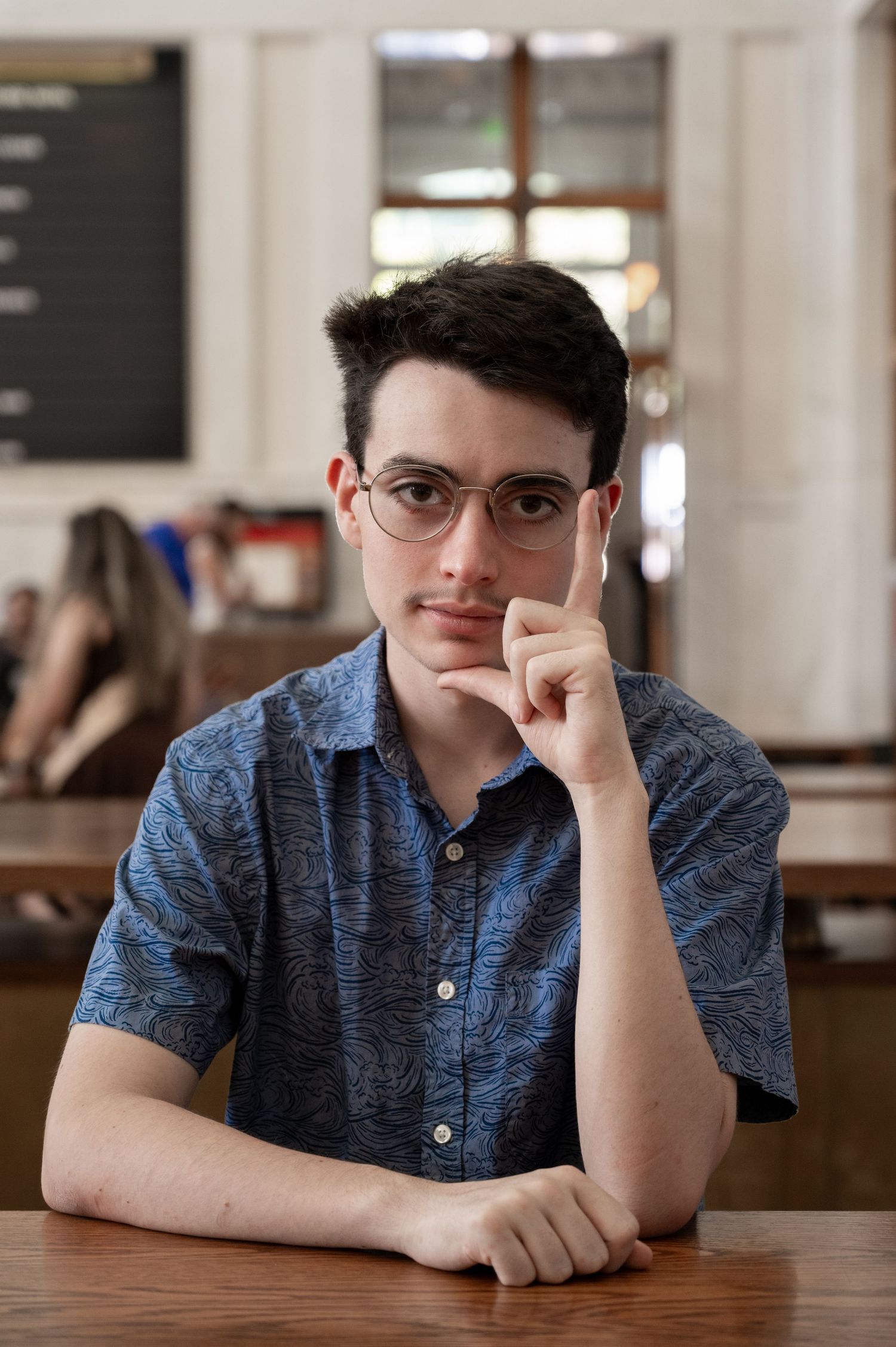 Young man with glasses sitting thoughtfully at a wooden table inside a cafe.