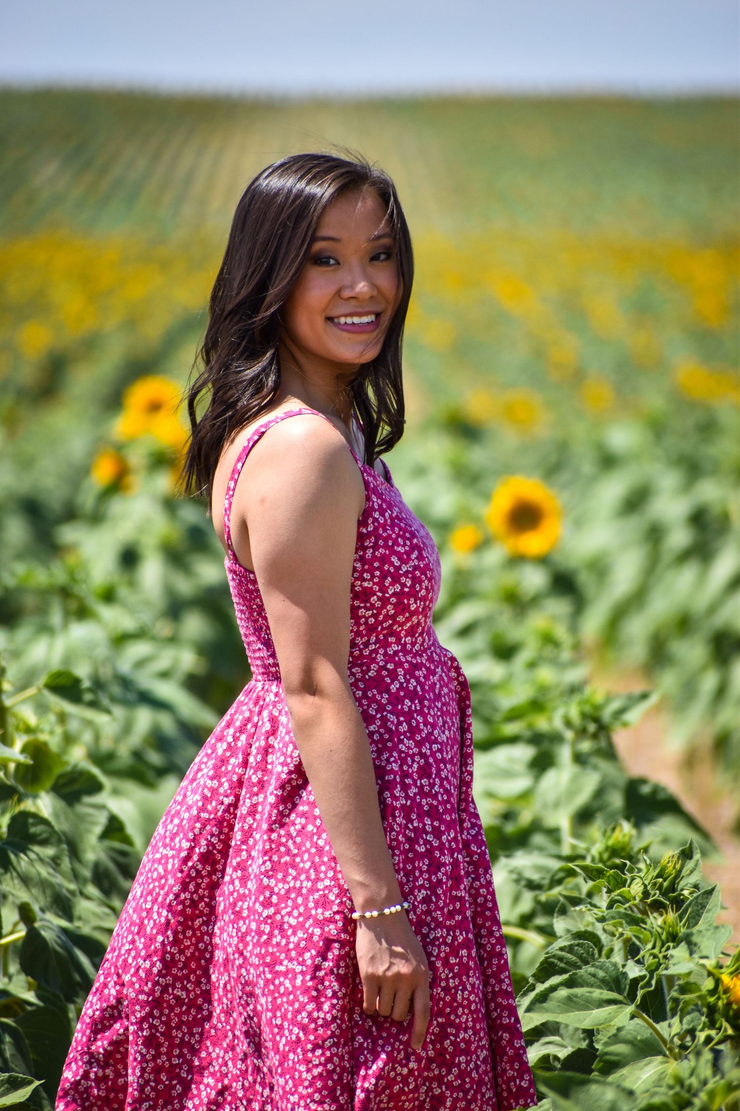Smiling young woman in pink floral dress stands in a vibrant sunflower field on a sunny day.