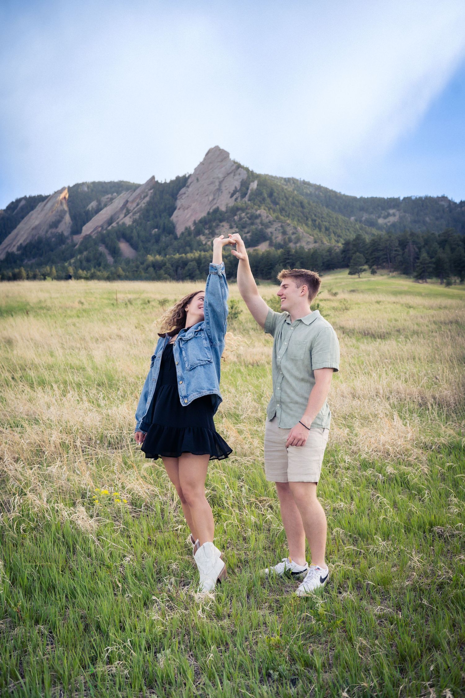 Happy couple dancing and spinning in a green meadow with dramatic rocky mountain peaks behind them.