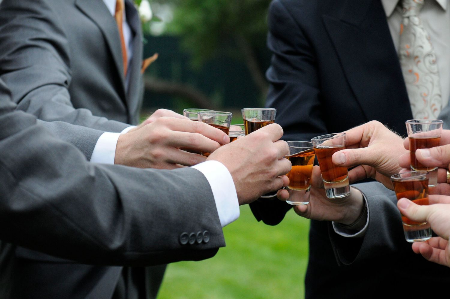 Wedding party members in suits clink shot glasses together in an outdoor toast celebration.