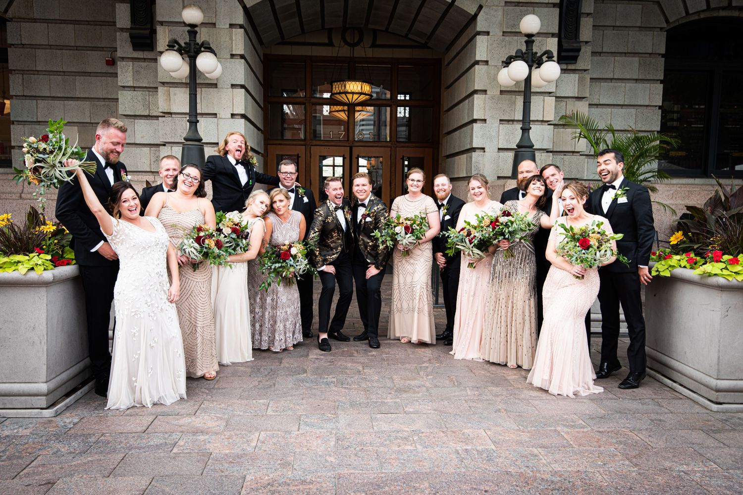 Wedding party poses outside elegant building with bridesmaids in champagne gowns and groomsmen in black tuxedos.