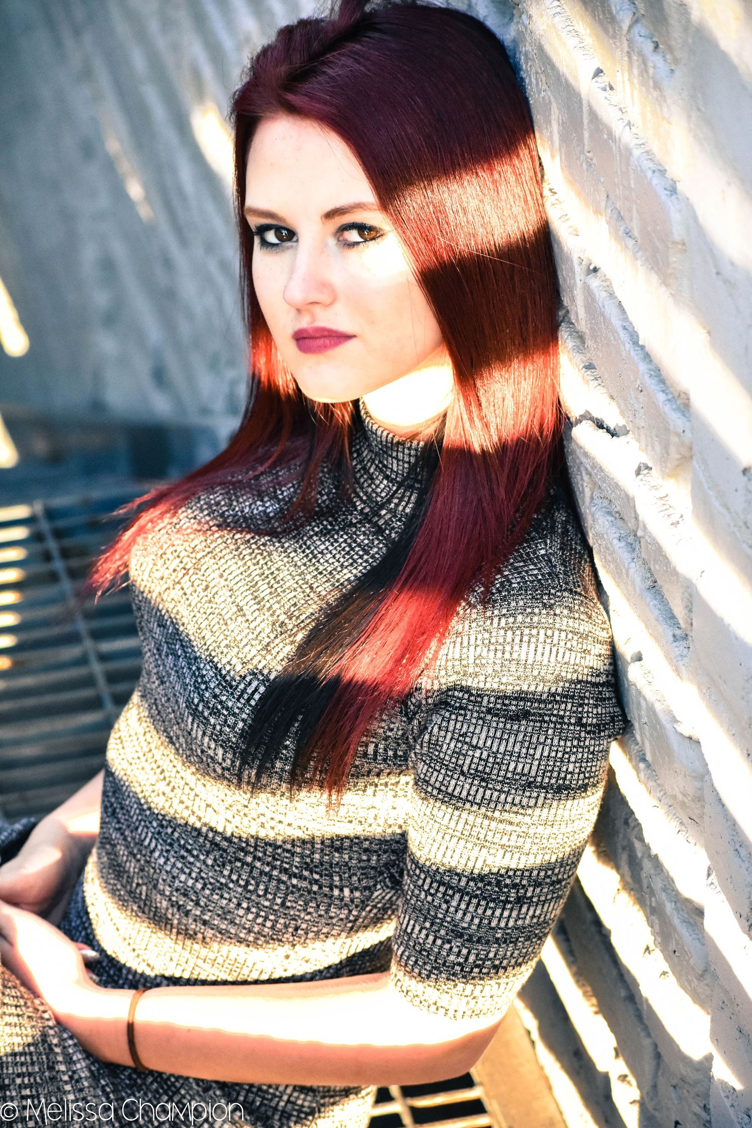 A young woman with long red hair in a striped knit sweater leans against a brick wall in natural sunlight.