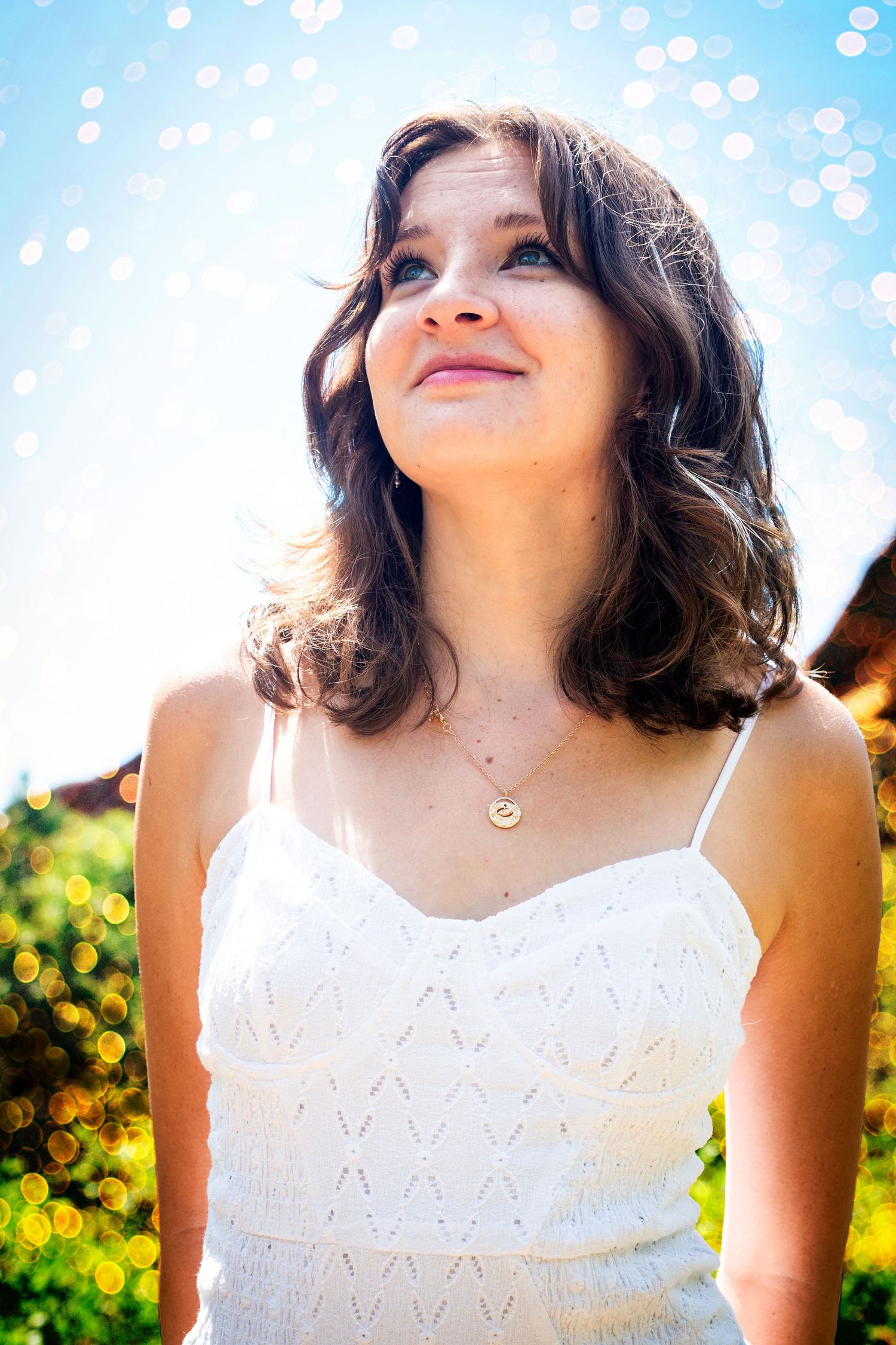 Teen girl in white sundress looks up smiling with bokeh sunlight effects in outdoor setting.