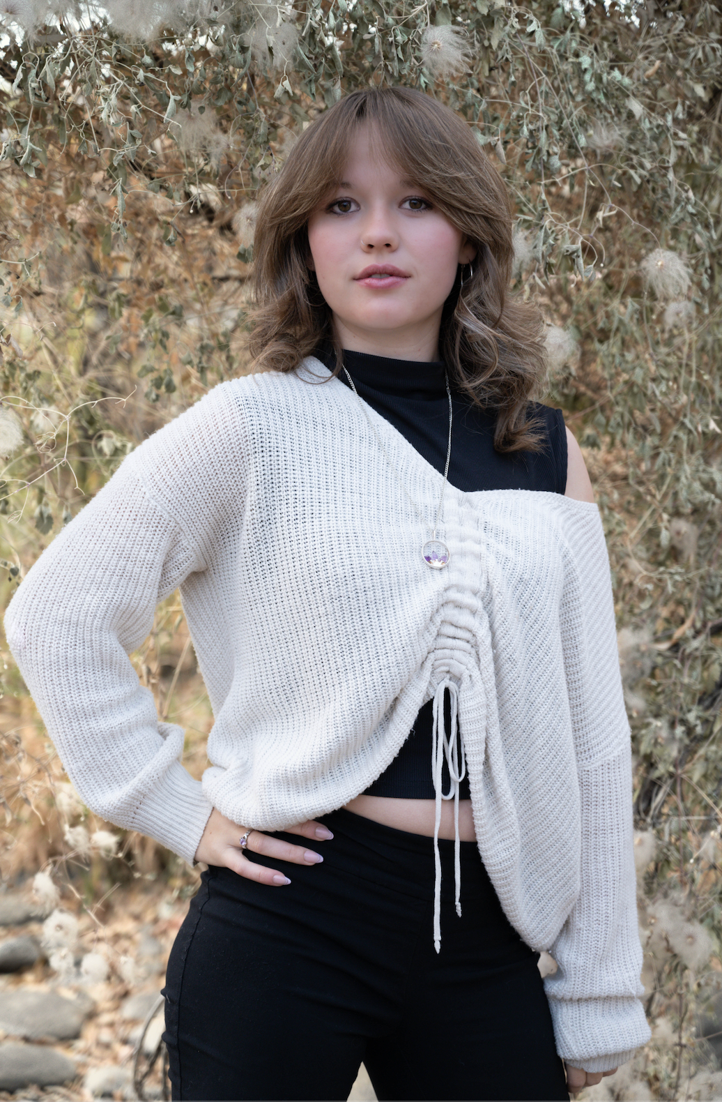 Teen girl in cream knit sweater and black top posing outdoors among dry foliage.