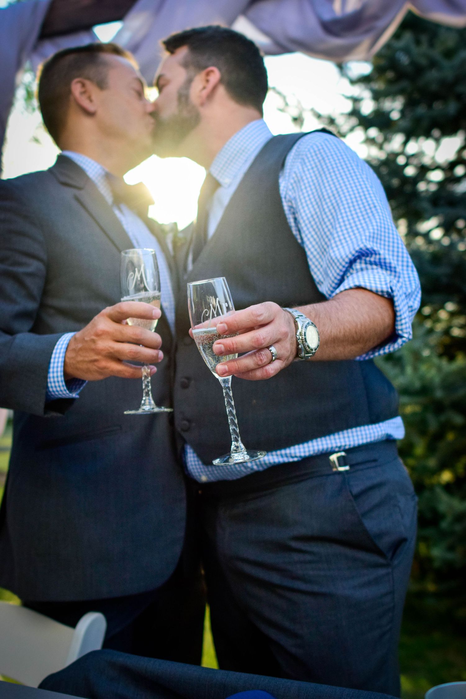 Two grooms kissing while holding engraved champagne flutes at their outdoor wedding ceremony at sunset.
