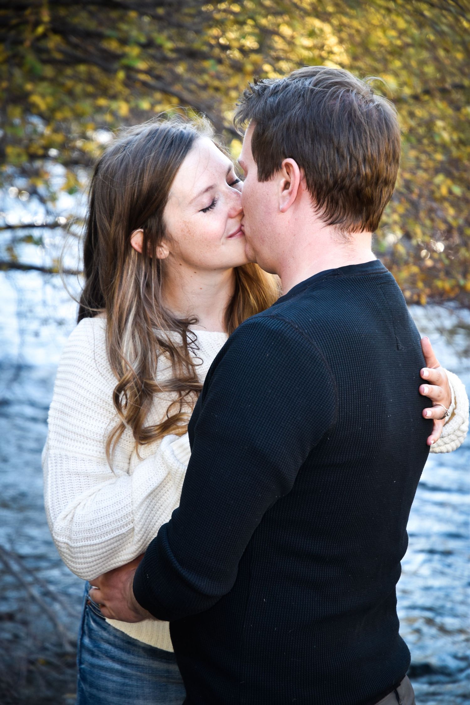 Couple sharing a tender kiss near a river surrounded by autumn foliage in natural setting.