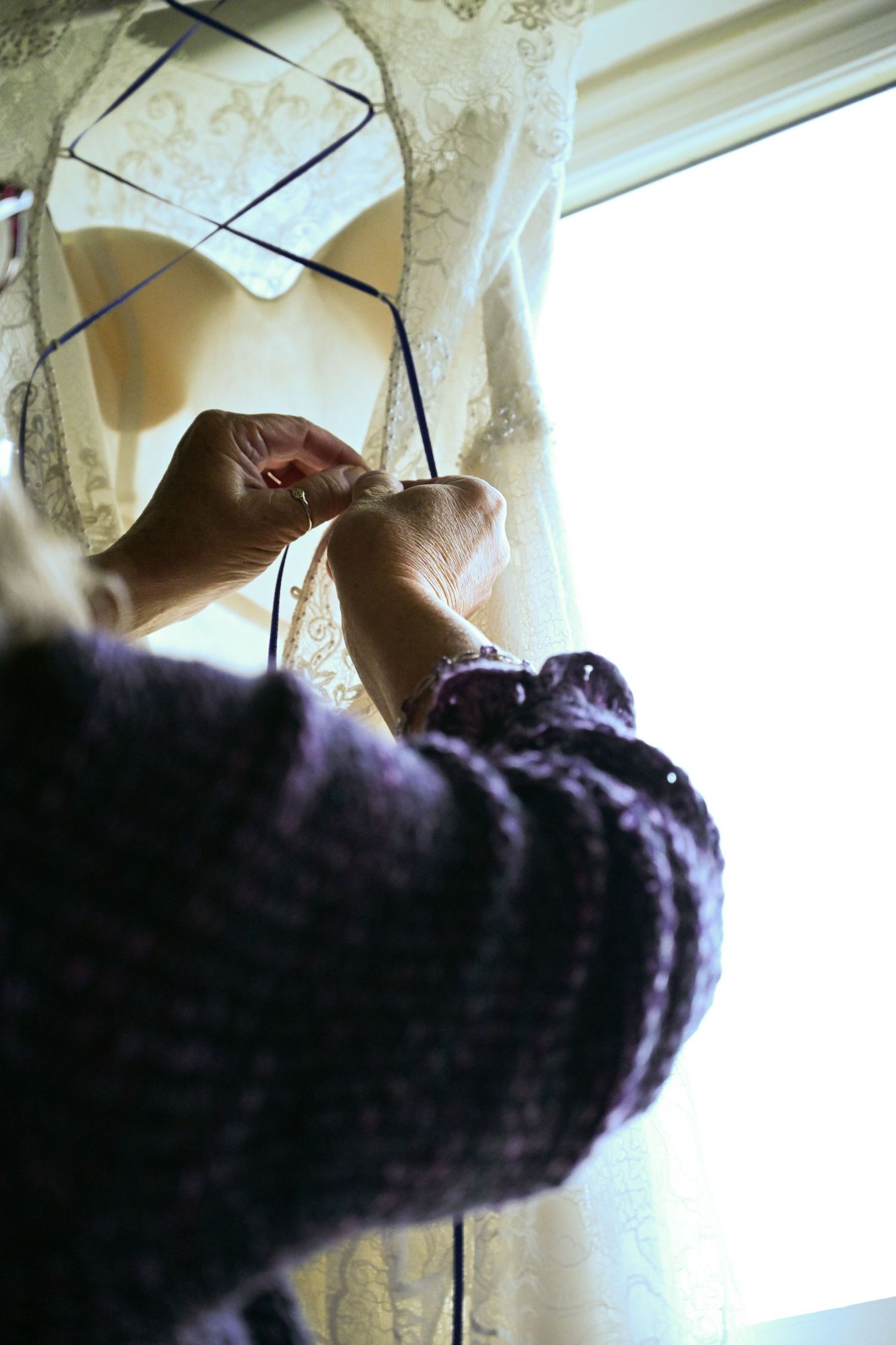 Close-up of hands carefully adjusting lace details on a wedding dress hanging by a window.