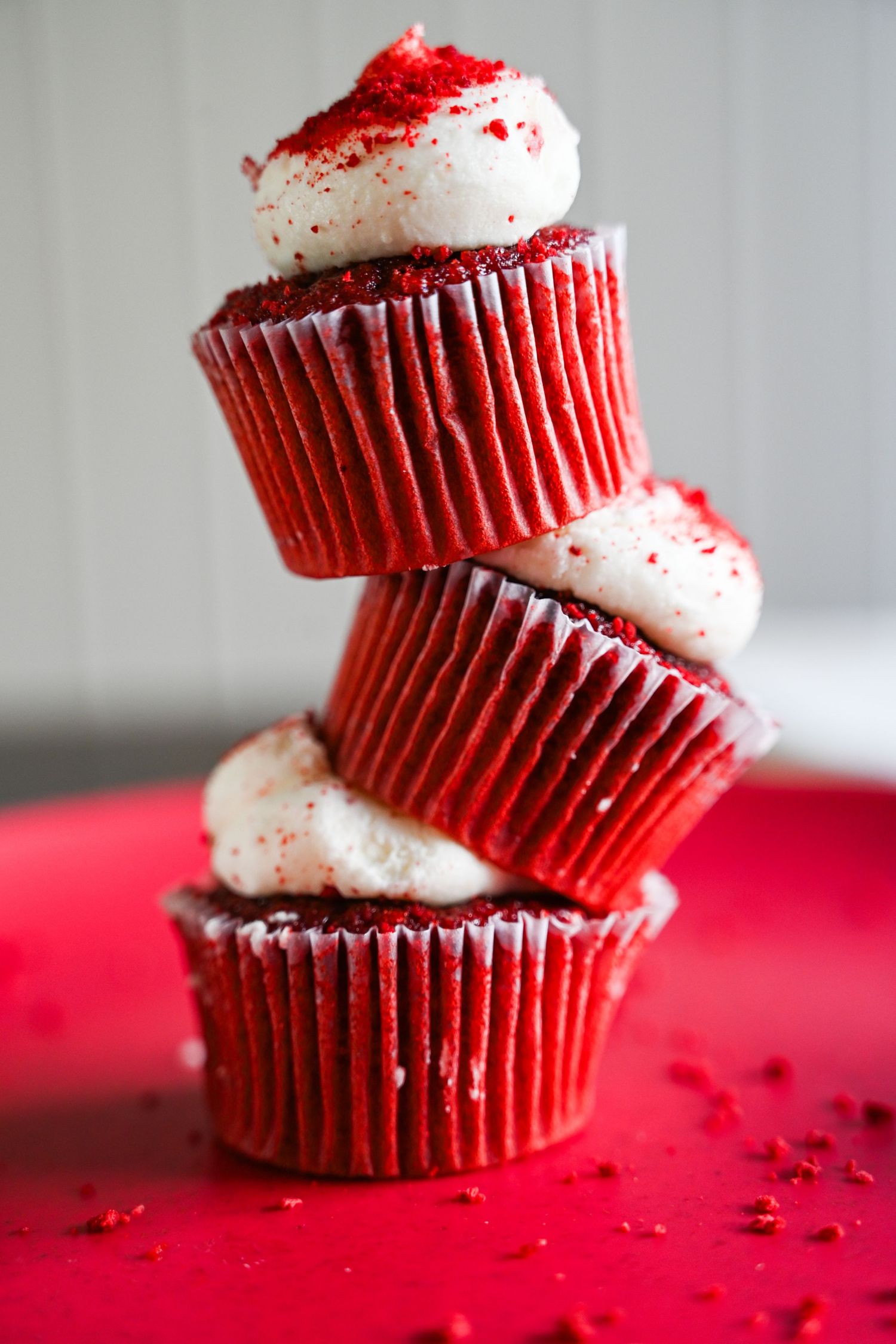 Three stacked red velvet cupcakes with white cream cheese frosting and red crumbs on a red surface.