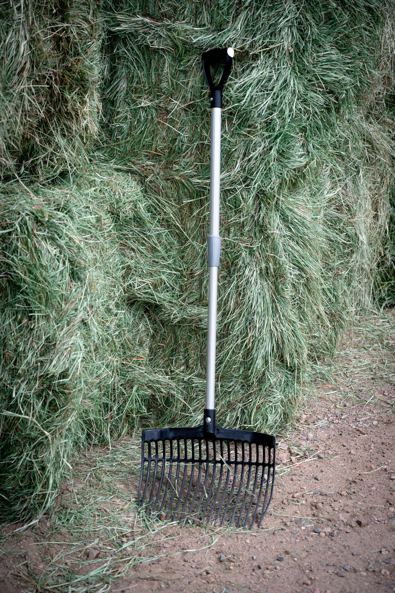 A black pitchfork leaning against stacked green hay bales on a dirt ground in a farm storage area.