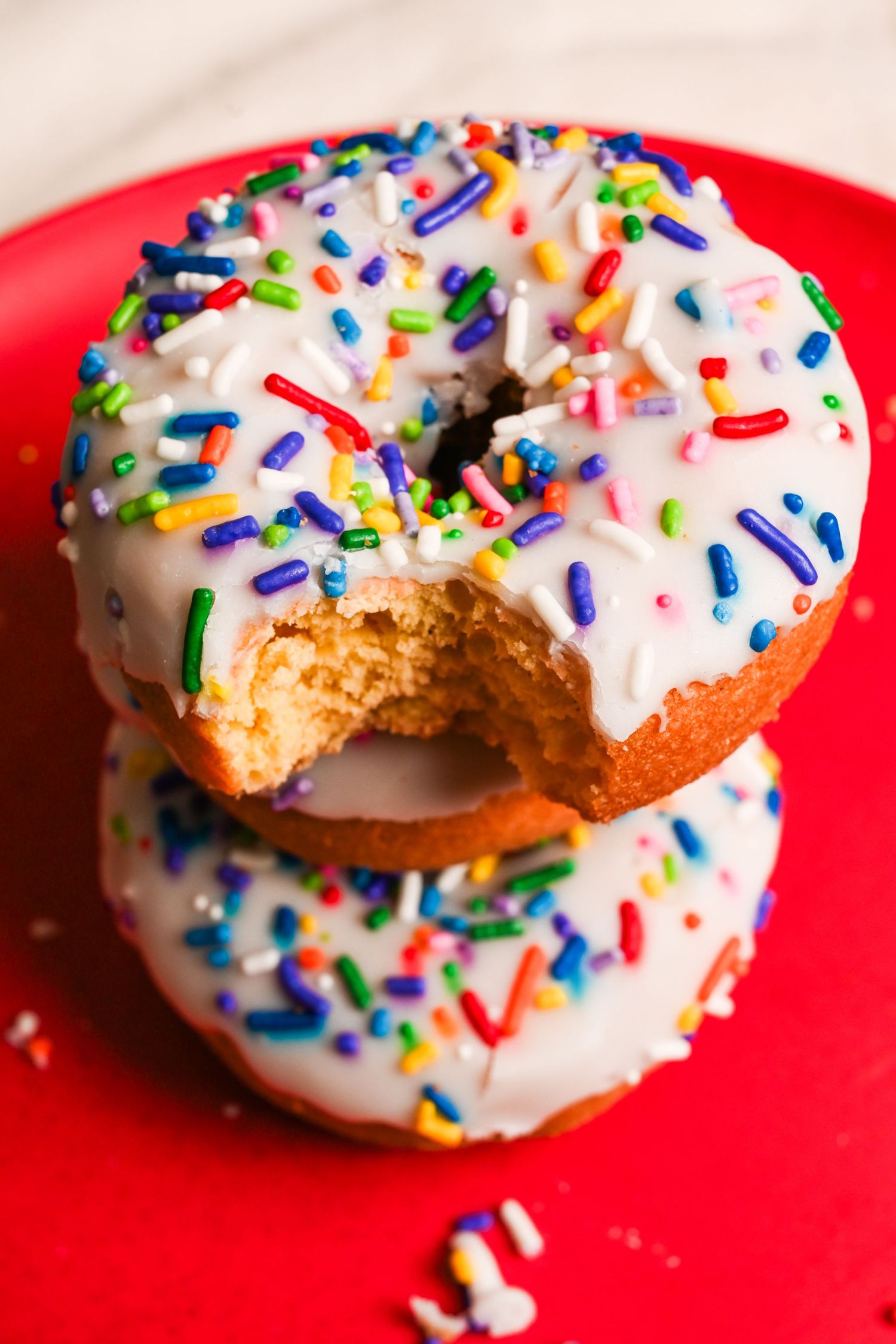 Two white-glazed donuts with rainbow sprinkles stacked on a red plate, one with a bite taken.