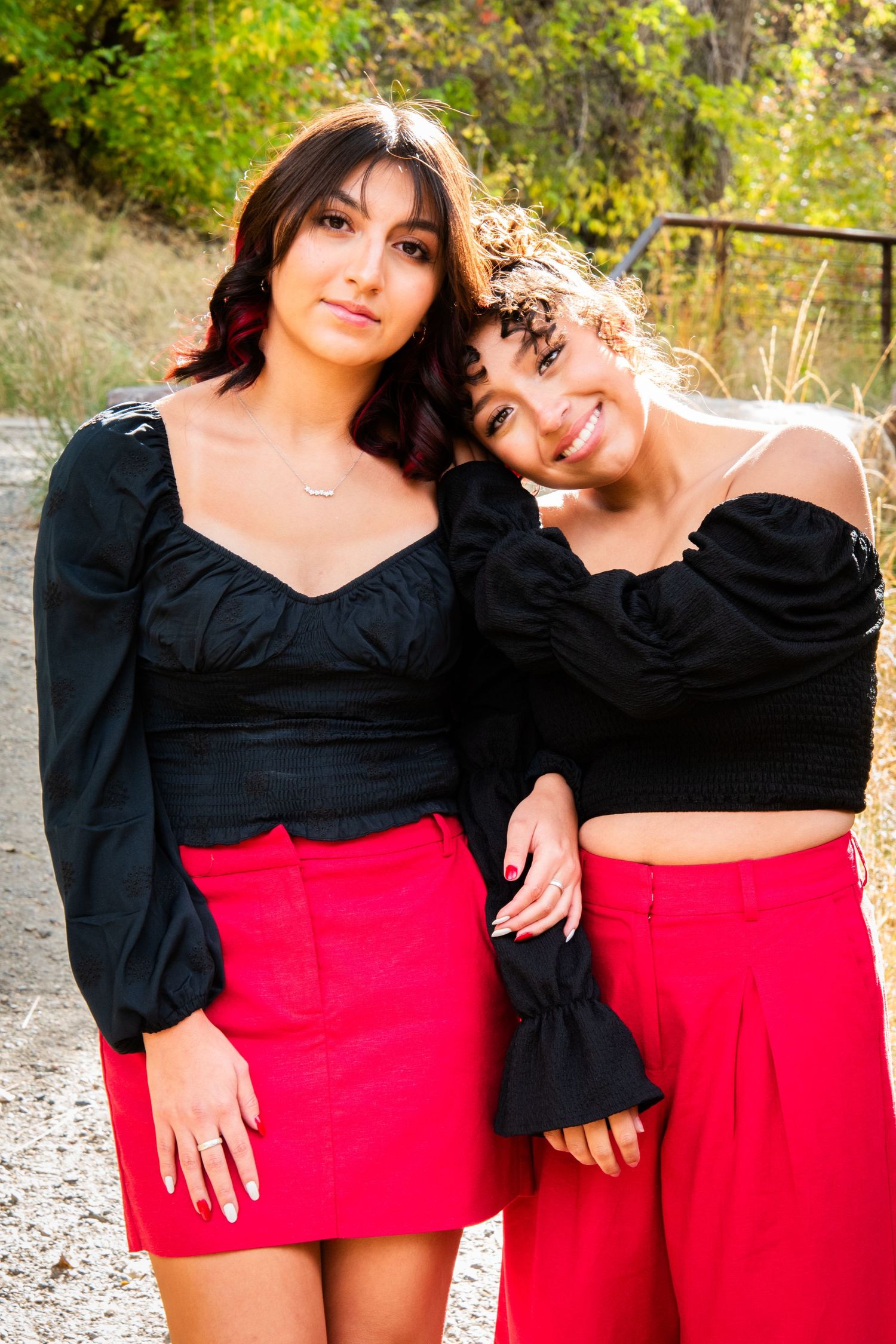 Two young women in matching black tops and red skirts pose together outdoors among green trees.