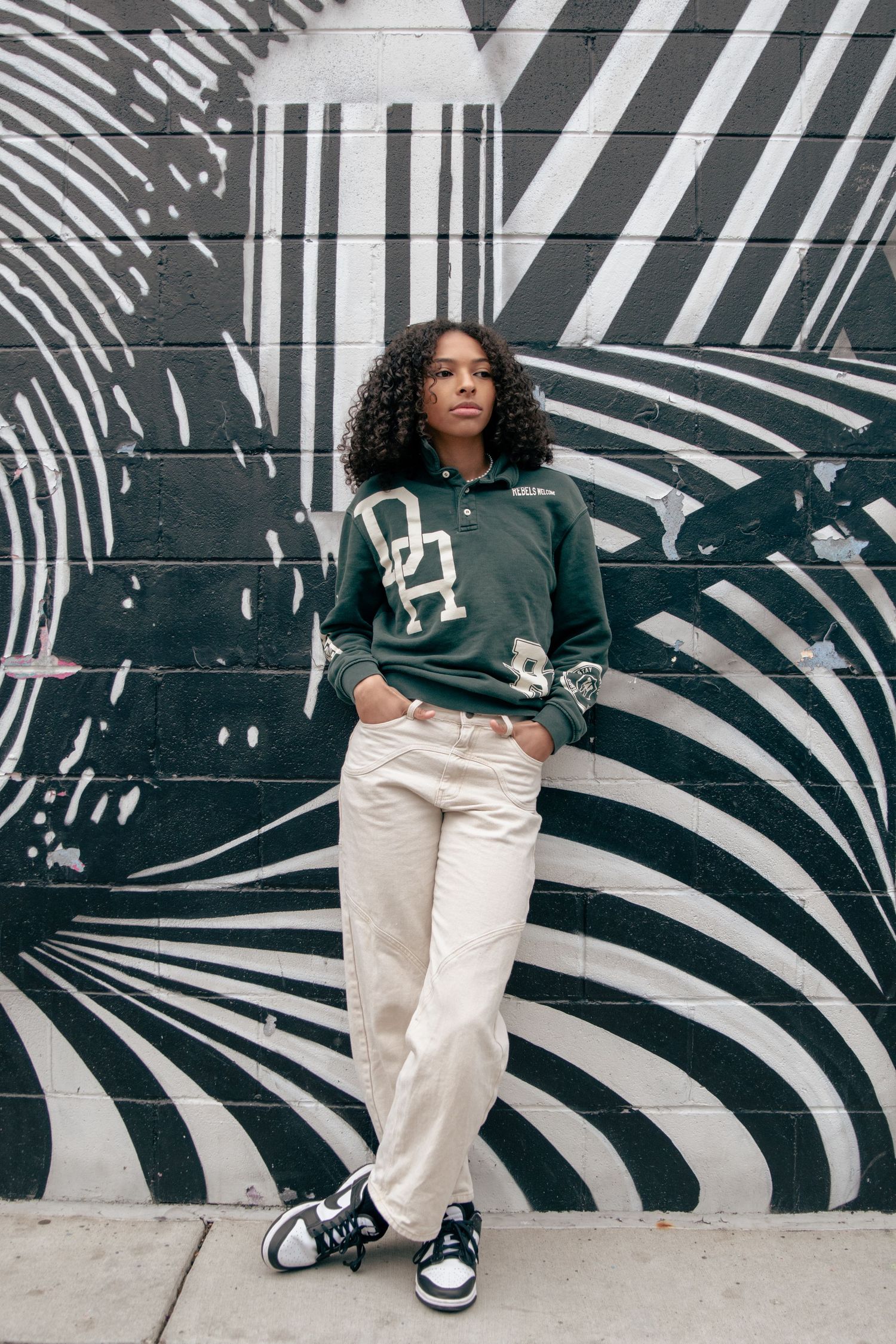Young woman in green varsity sweatshirt and cream pants stands before a black and white mural.