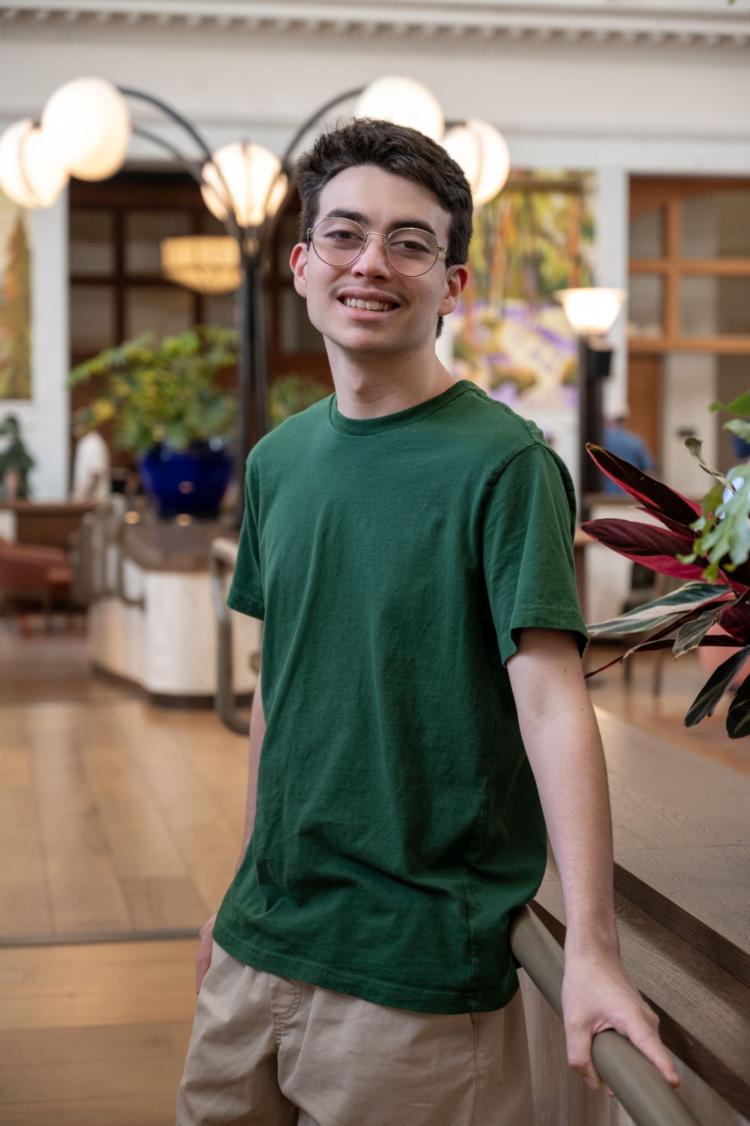 Smiling teen boy in green t-shirt standing in bright indoor atrium with plants.