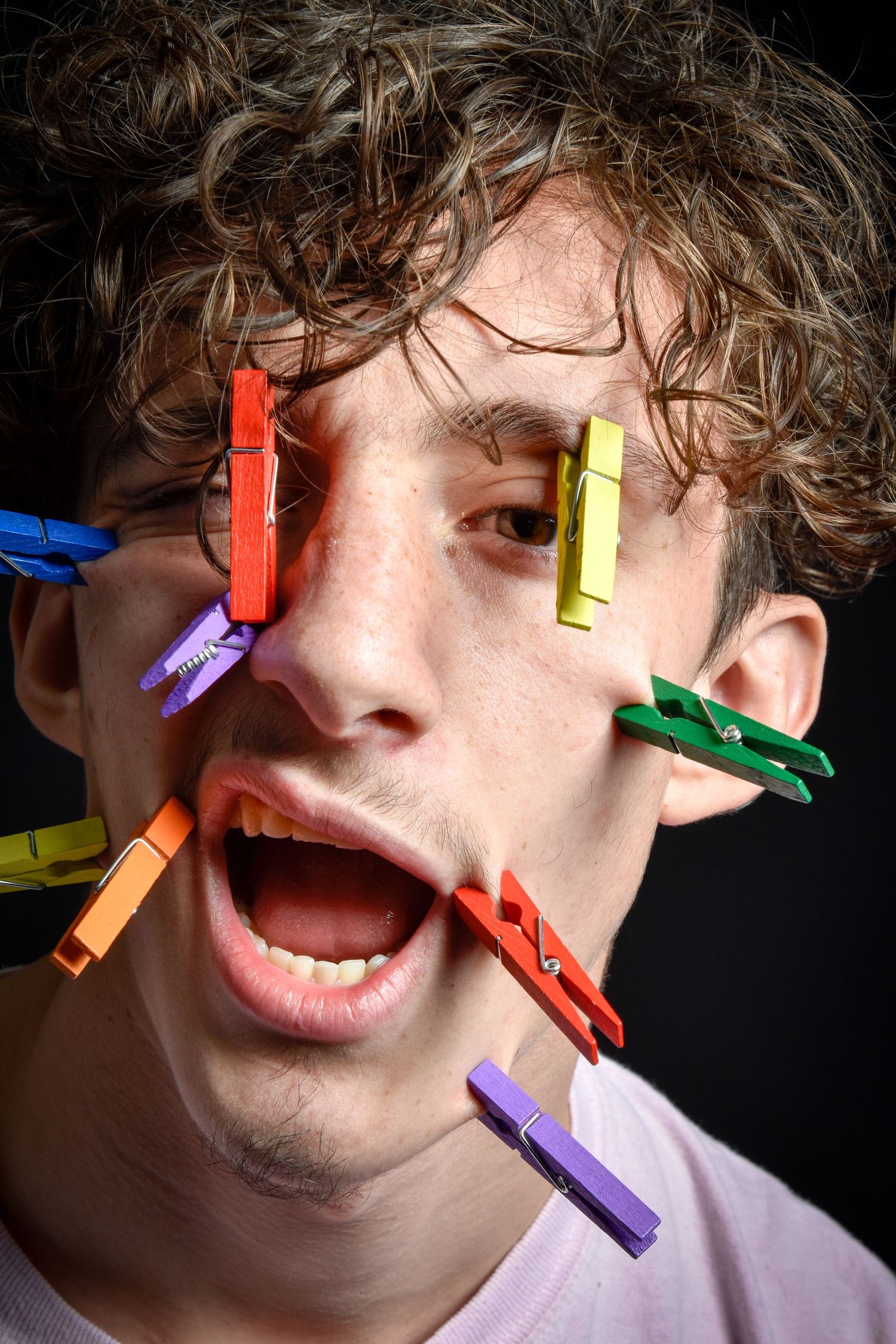 Young man with curly hair and colorful clothespins clipped to his face, mouth open wide, against dark background.