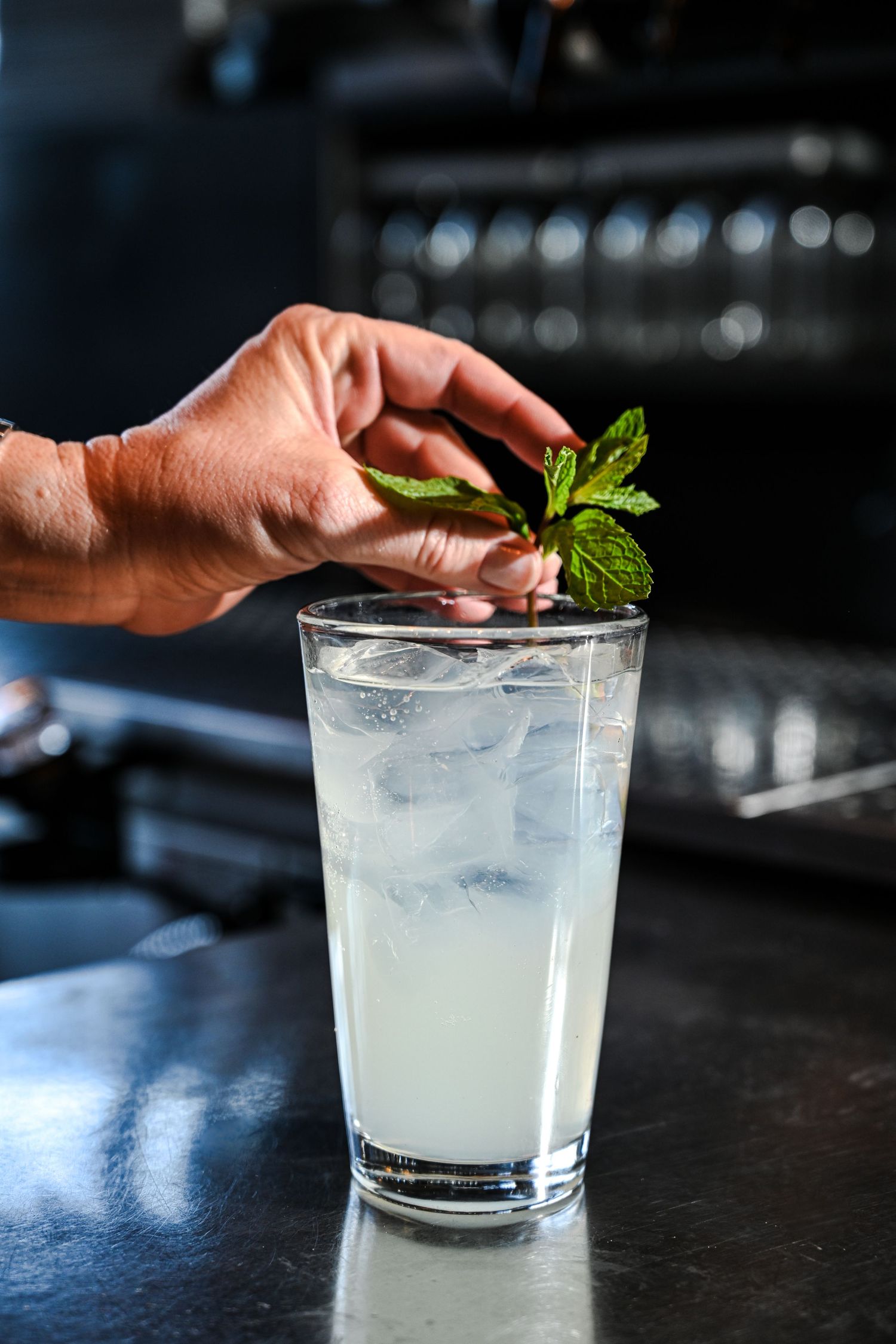 Bartender garnishing a tall iced lemonade cocktail with fresh mint leaves at a bar.